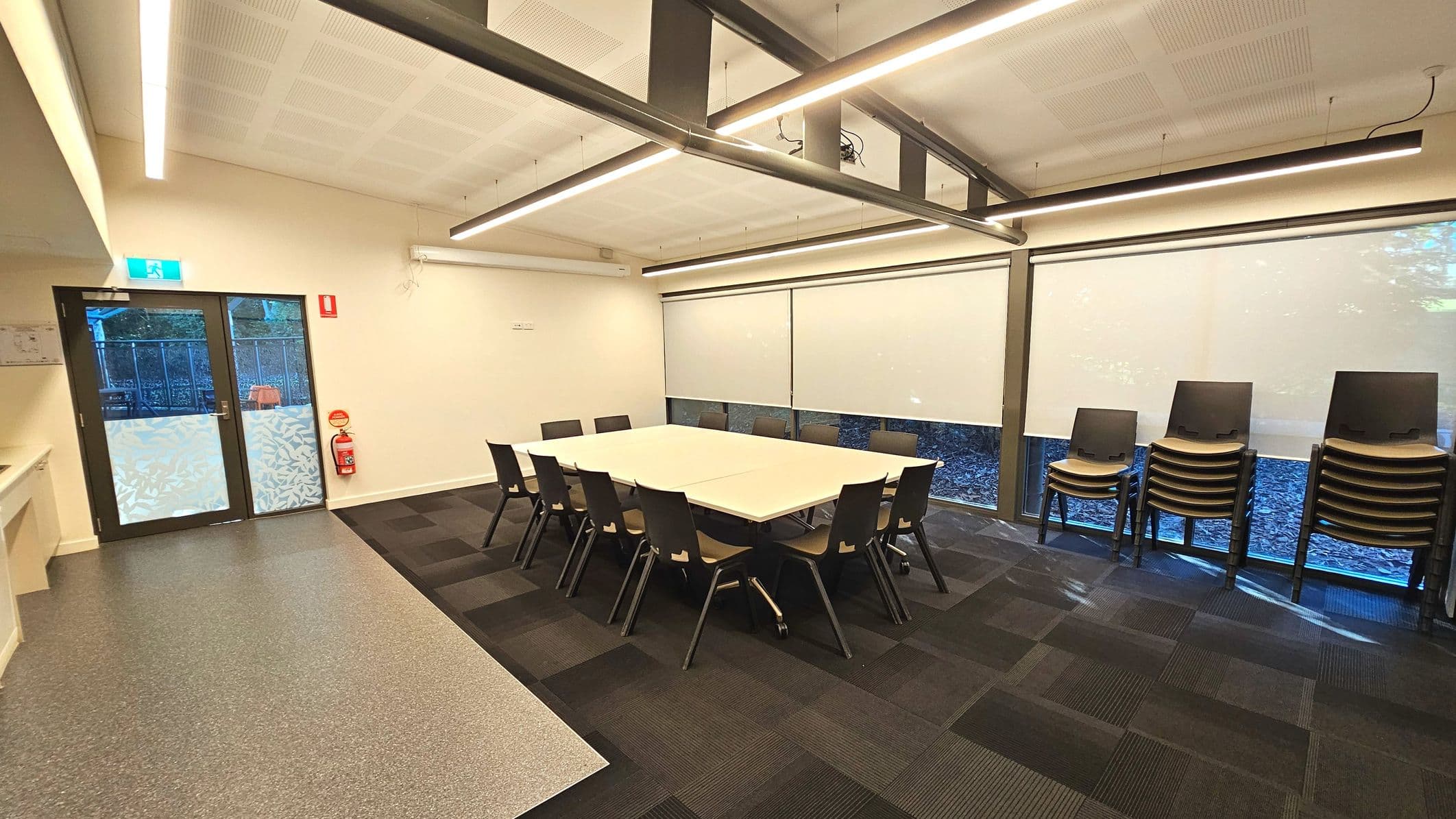 A modern meeting room with a large square table surrounded by twelve black chairs. The room features a mix of grey speckled and dark carpeted flooring. Large windows with roller blinds line one wall, and a stack of additional chairs is placed nearby. A glass door with a patterned lower half is on the left, next to a fire extinguisher. The ceiling has linear lights and exposed beams, creating an open feel.
