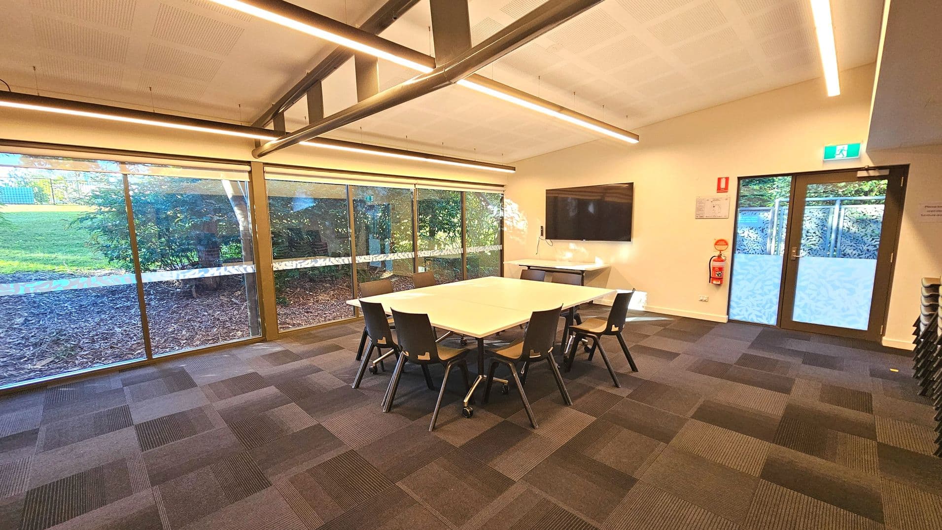 A modern meeting room with a large rectangular table surrounded by eight black chairs on a patterned carpeted floor. The room features a wall-mounted flat-screen TV and a fire extinguisher near a glass door with frosted patterns. Large windows on one side offer a view of greenery outside. The ceiling has exposed beams and linear lights, creating a spacious feel. An emergency exit sign is visible above the door.