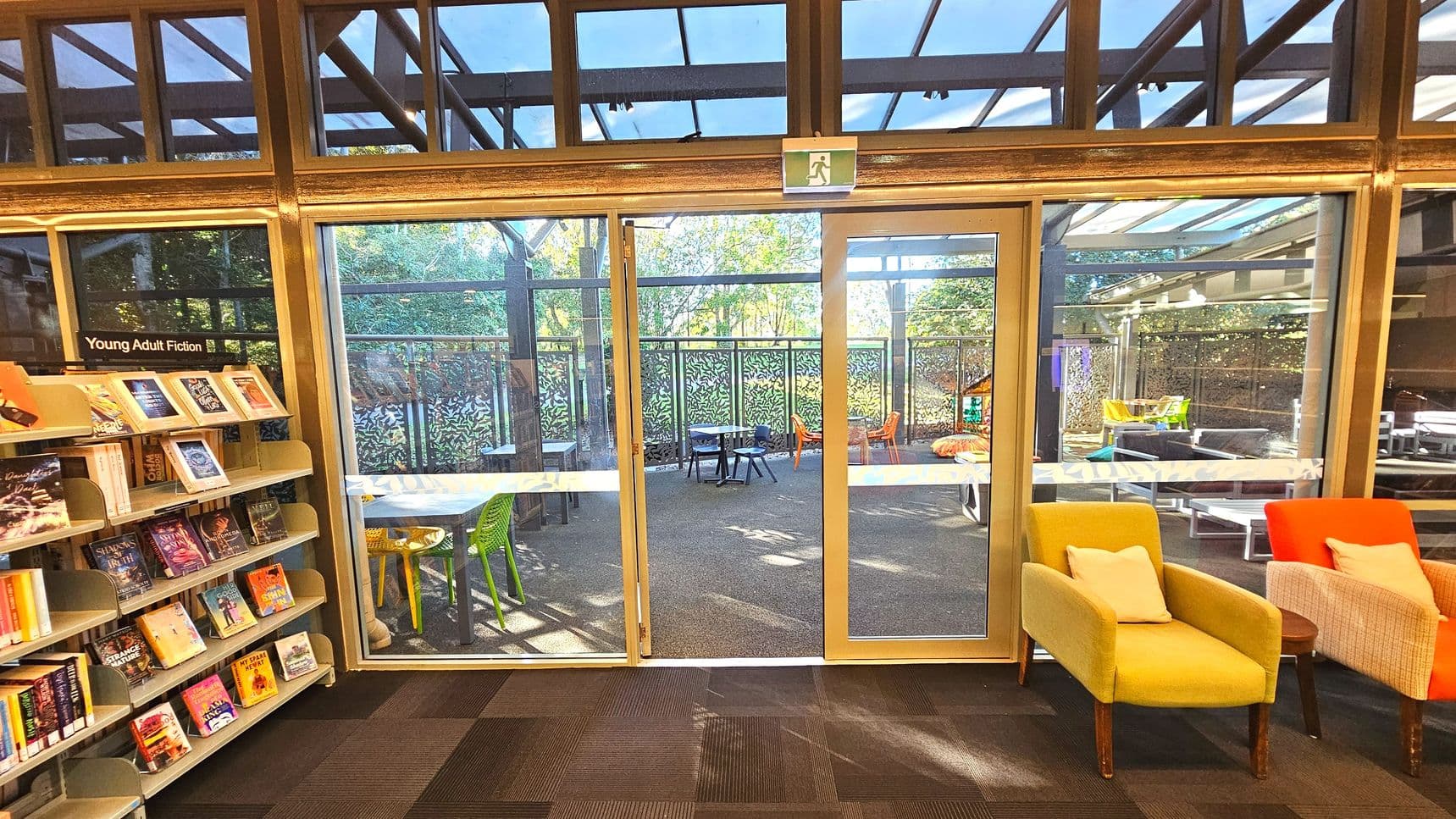 A library interior with a focus on a glass door leading to an outdoor area. To the left, a bookshelf labelled 'Young Adult Fiction' is filled with books. The door has a green exit sign above it and opens to a patio with colourful chairs and tables. The floor inside is carpeted in a dark pattern, while the patio has a textured surface. Brightly coloured armchairs with cushions are positioned near the door.
