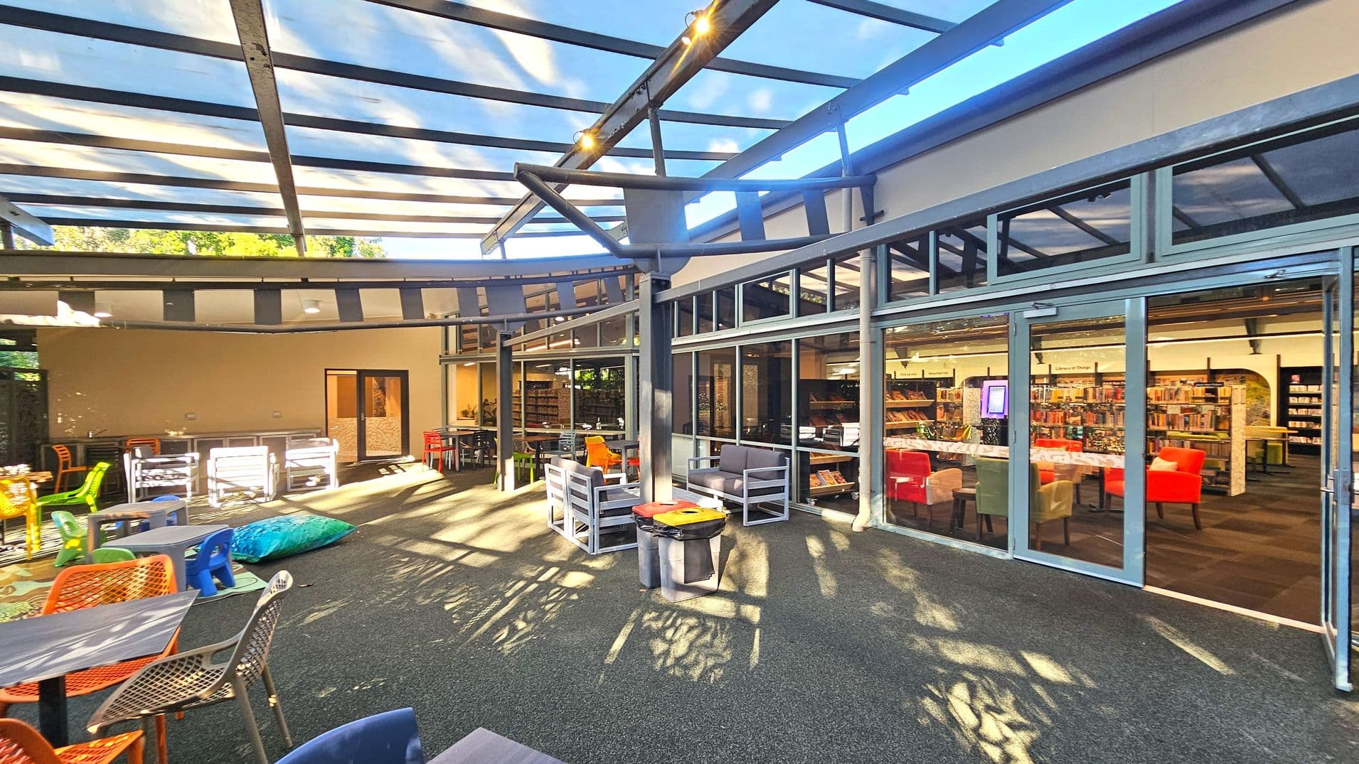 An outdoor seating area with a transparent roof made of glass panels supported by metal beams. The space features various colourful chairs and tables, including orange and green chairs, and a blue bean bag. Adjacent to this area is a building with large glass doors and windows, revealing an interior filled with bookshelves and red and green chairs. The floor is covered with dark grey carpet-like material.