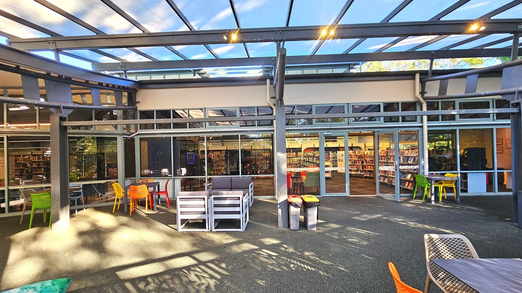 An outdoor seating area with colourful chairs and tables is adjacent to a building with large glass windows and doors. Inside, bookshelves are visible, suggesting a library or bookstore. The area is covered by a transparent roof with metal beams, allowing natural light. The ground is paved with a textured surface, and there are three bins near the entrance. The space feels open due to the glass walls and roof.