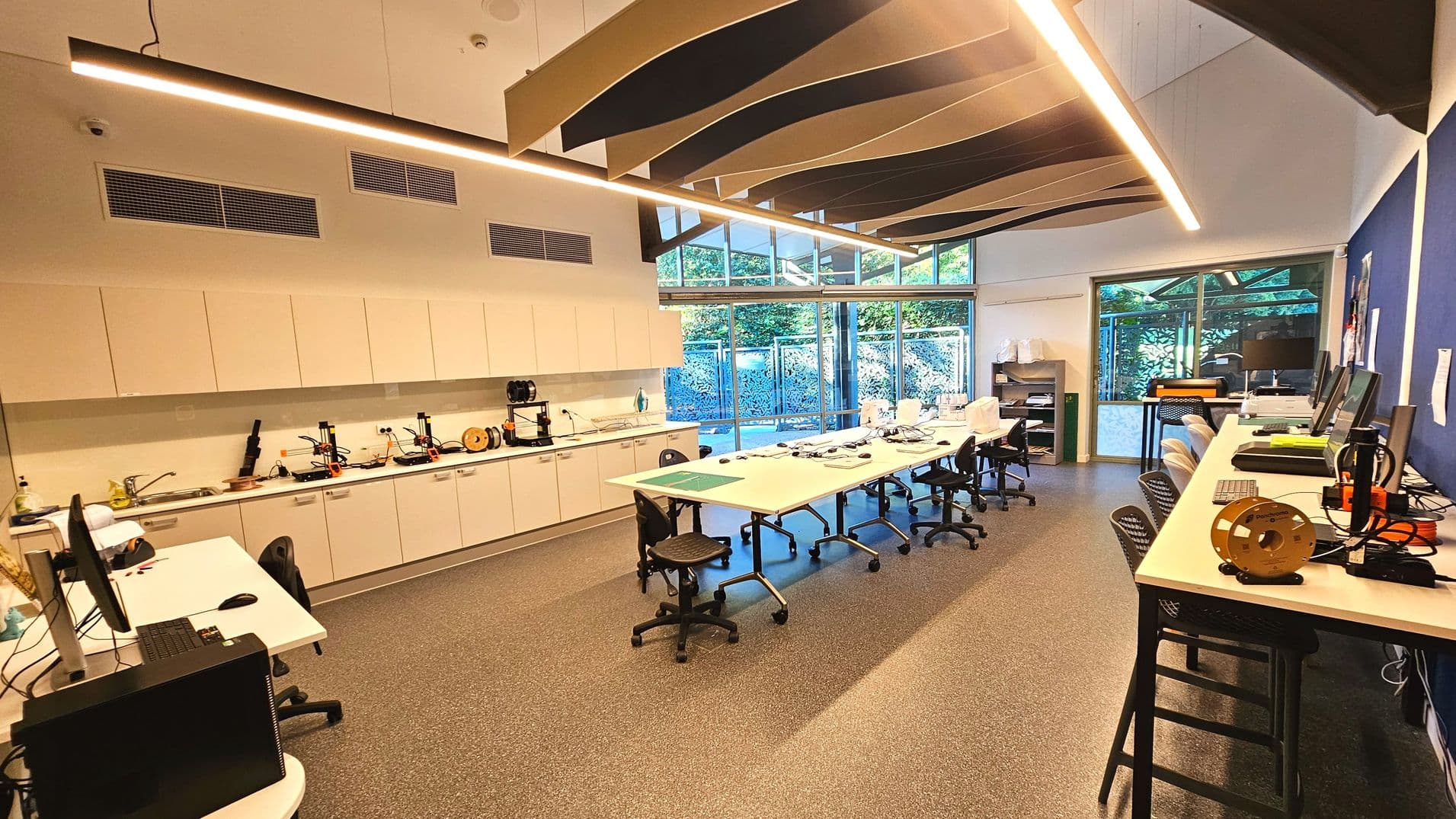 A modern laboratory with a long central table surrounded by swivel chairs, equipped with computers and various electronic devices. The room features a high ceiling with wave-like panels and large windows along one wall, allowing natural light to illuminate the space. Cabinets line another wall, housing equipment and supplies. The floor is covered with speckled grey tiles, and there are additional workstations along the perimeter.