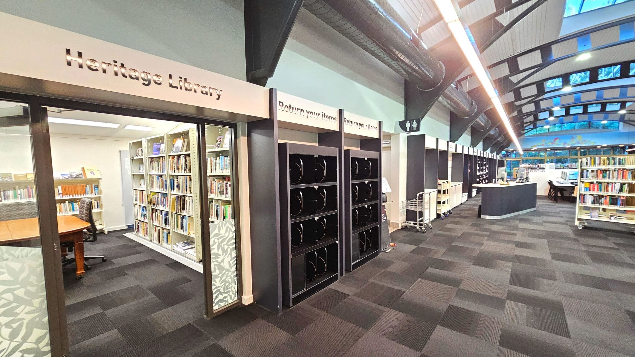 A library interior featuring a 'Heritage Library' section on the left with glass doors and bookshelves inside. Adjacent are black return slots labelled 'Return your items'. The space has a high, curved ceiling with exposed beams and skylights. The floor is covered with patterned carpet tiles. A curved desk is visible further in, with bookshelves and a sign for printing nearby. A restroom sign is mounted on the wall.