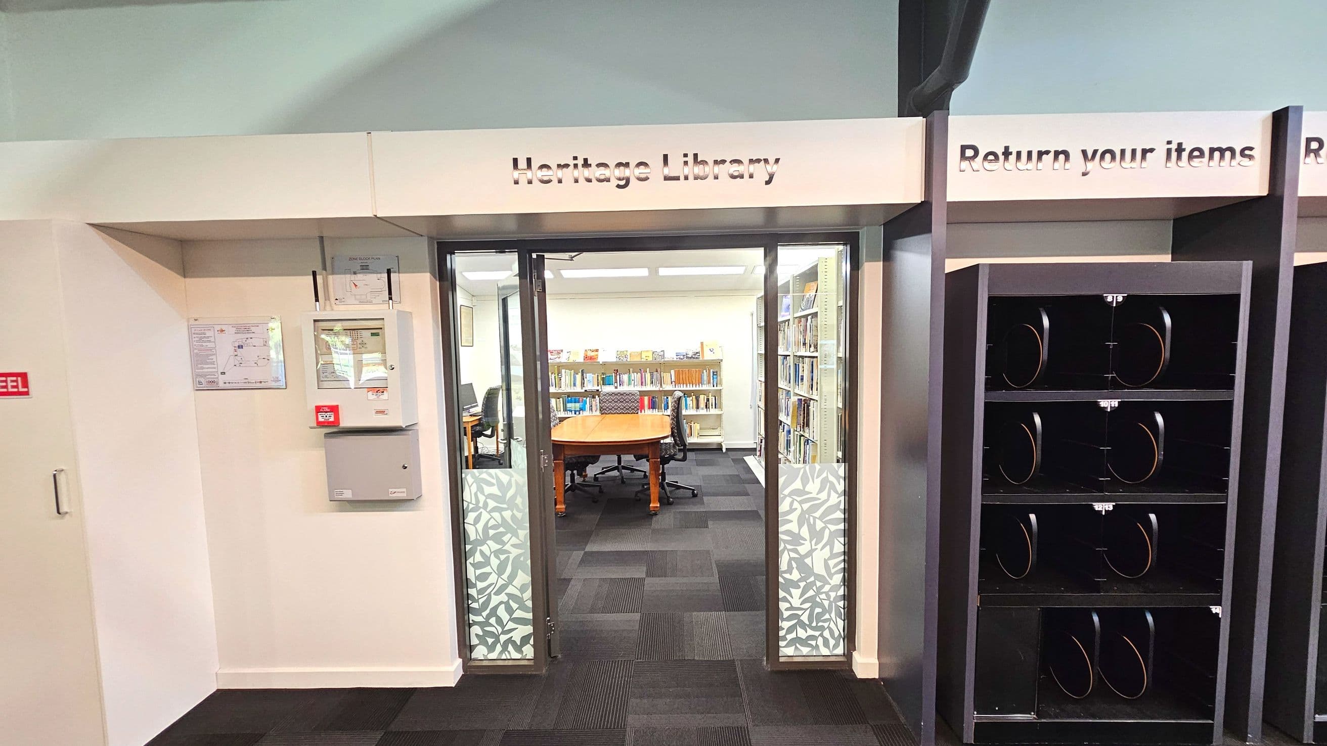 The image shows the entrance to a Heritage Library, marked by a sign above glass doors with frosted leaf patterns. Inside, a wooden table and chairs are visible, surrounded by bookshelves filled with books. To the right, there is a 'Return your items' sign above empty shelving units. The floor is covered with dark carpet tiles, and the space feels enclosed by walls and shelving.