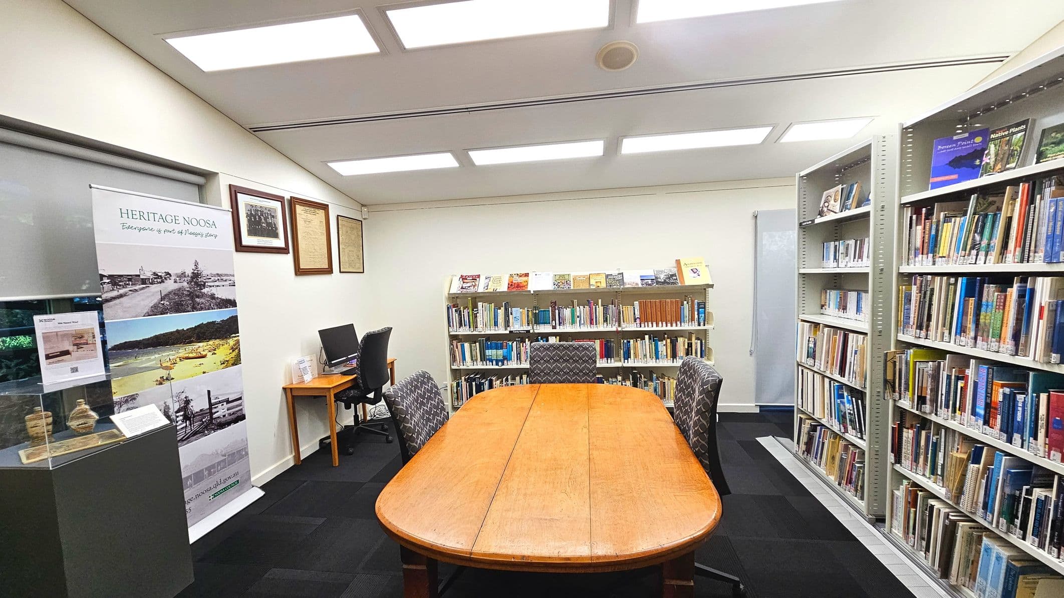 A library room with a central wooden table surrounded by four patterned chairs. Bookshelves line the right wall, filled with books. A smaller bookshelf is against the back wall, displaying more books and magazines. On the left, a computer desk with a chair is adjacent to a display case and a 'Heritage Noosa' banner. The room is well-lit with ceiling lights and has a black carpeted floor.