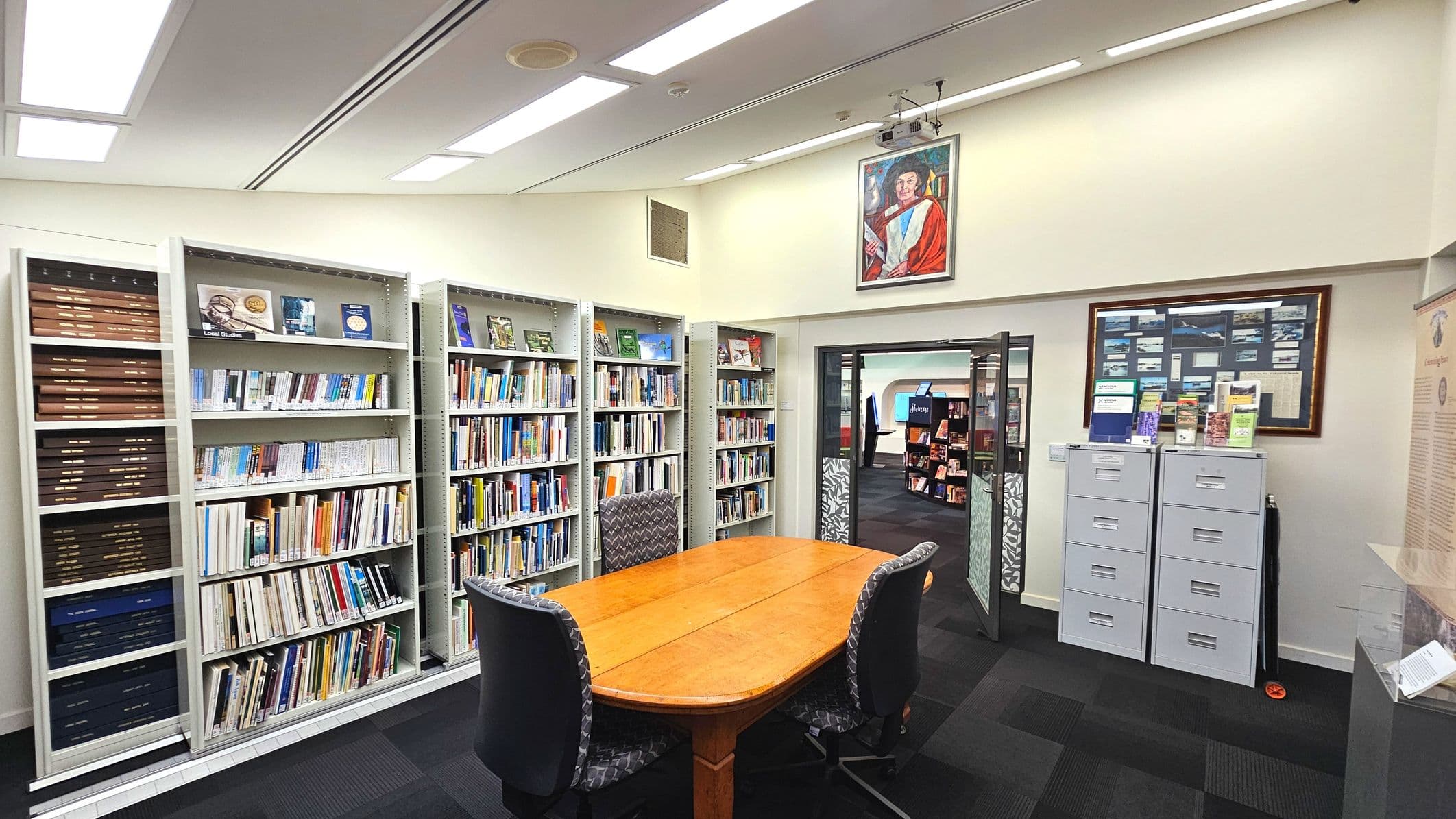 A library room with three tall white bookshelves filled with books and some decorative items. In the centre, a wooden oval table is surrounded by four cushioned chairs. To the right, two grey filing cabinets are topped with brochures and a framed collage. A glass door leads to another room with more bookshelves. The floor is carpeted in dark tiles, and a portrait hangs above the door.