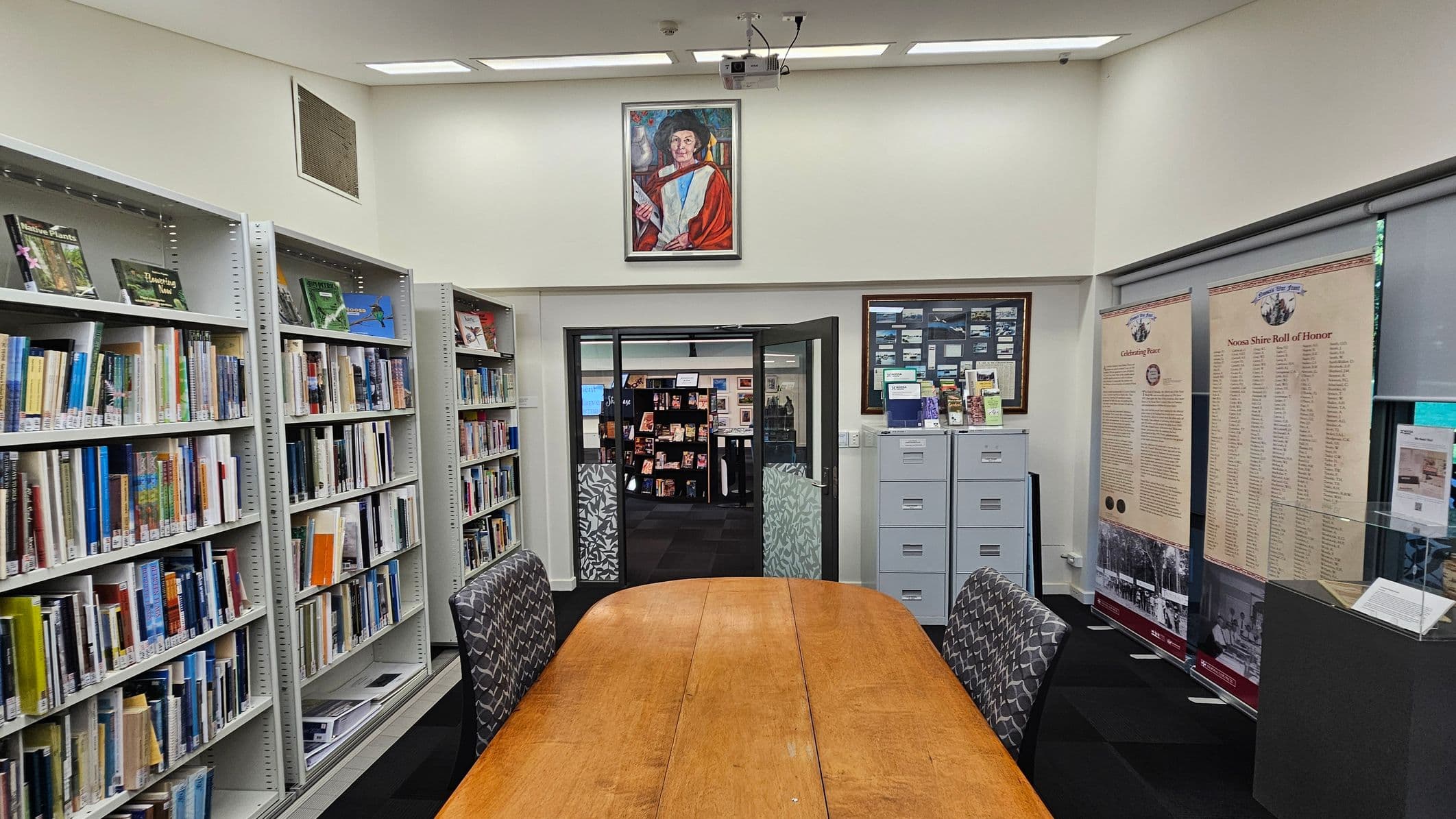 A library room with tall white bookshelves filled with books on the left. A long wooden table with patterned chairs is in the centre. Above the doorway is a portrait of a person in academic robes. The doorway leads to another room with displays. On the right, there are informational banners and a glass display case. The floor is carpeted, and the ceiling has recessed lighting.