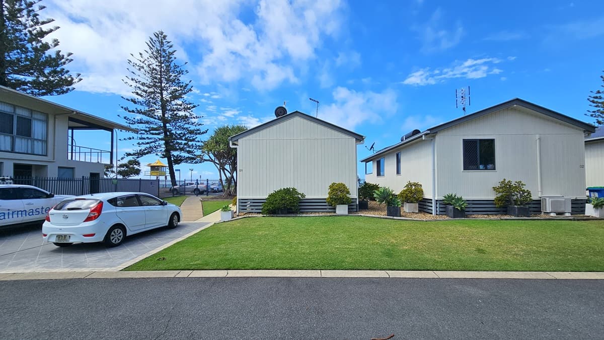 To the right, two low-set buildings with pitched roofs and white facades stand amid plants in planter boxes and green grass. To the left, a two-storey building with a veranda is accompanied by a car park with two vehicles. In the foreground, a bitumen road is visible, while in the background, the ocean and pine trees stretch out. A watchtower is also visible.
