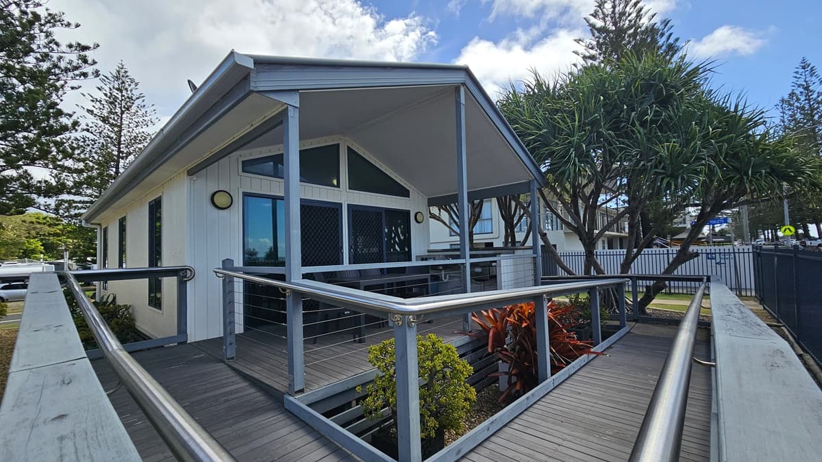 A low-set building with a white facade stands amidst plants and green grass. A ramp with handrails extends from the car park to the veranda, where tables and chairs are arranged. The ramp transitions from concrete to wooden planks. Trees and grass are visible in the background. On the right is a metal fence securing the area.