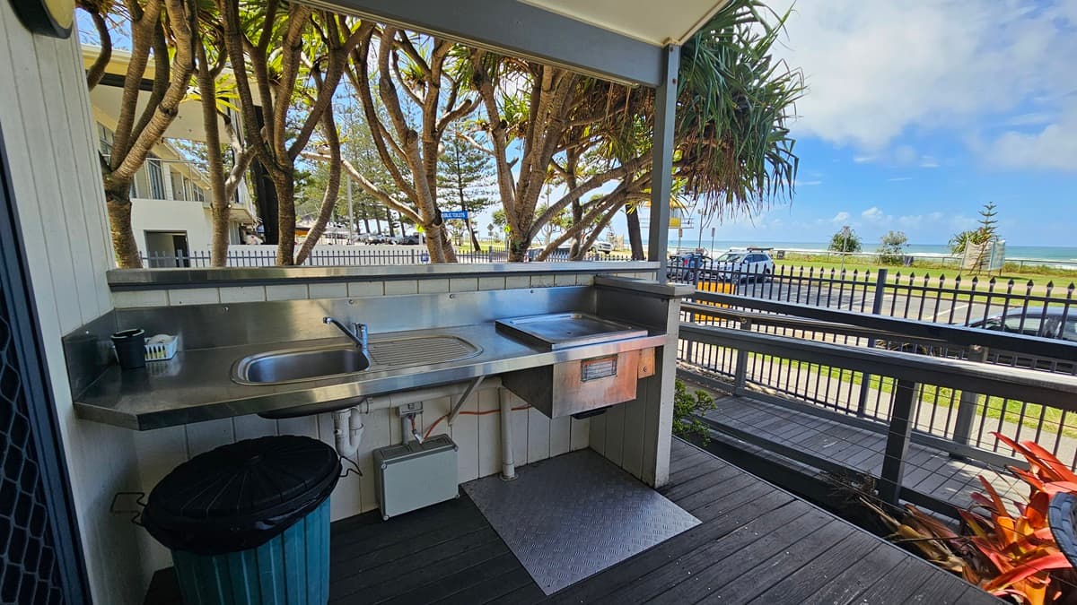The veranda has a barbecue and a sink, with a rubbish bin placed underneath. To the left is a glass door entrance to the cabin, equipped with a mesh door. The floor has wooden planks, and the ceiling and walls are white with light fixtures. On the right, a ramp with handrails is present. A low fence secures the veranda, while plants and trees adorn the front.
