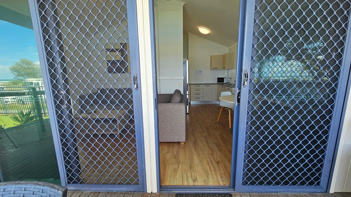 Entrance to the cabin is through a manual glass sliding door equipped with a mesh screen. The door is open, offering a view inside. The interior features a timber floor, with visible elements including the kitchen, a couch, and a dining table.