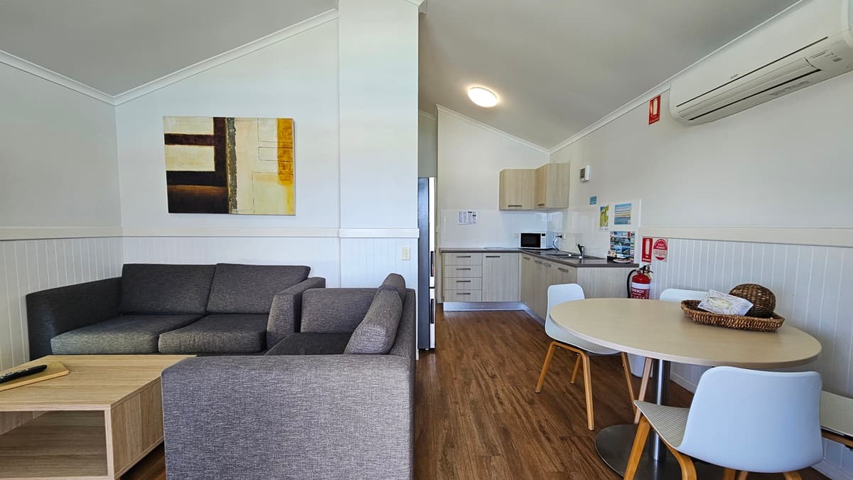 Inside the cabin, a grey couch and coffee table are set to the left, while a round table with chairs is positioned to the right. Adjacent to the table is the kitchen area, featuring a sink, kitchen cabinets, oven, and fridge. The wall is painted white with a framed decoration, and an air conditioner and fire extinguisher are mounted on it. The ceiling is white, and the floor is timber.