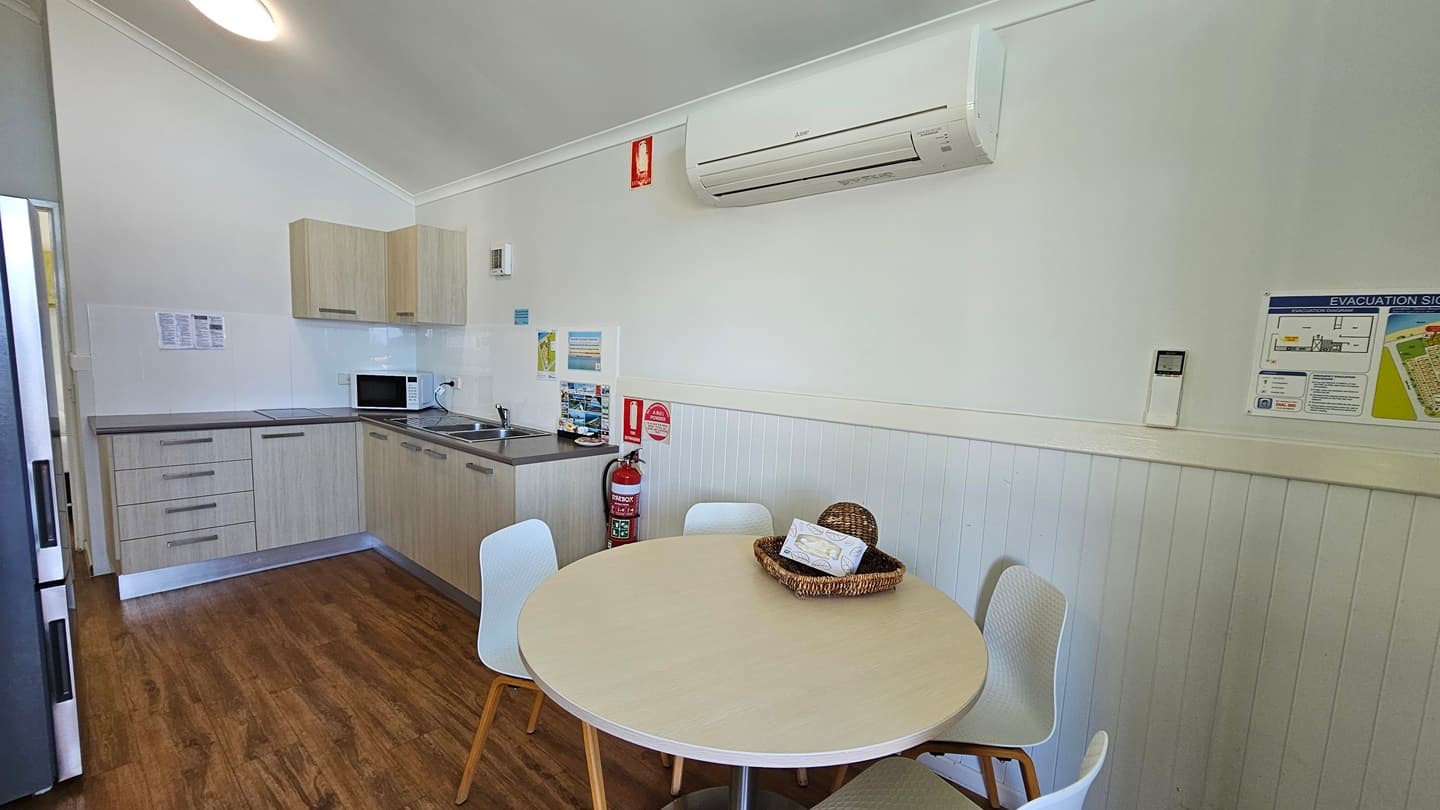 A round table with chairs is positioned to the right. Decoration and a tissue box sit on top. Adjacent to the table is the kitchen area, featuring a sink, kitchen cabinets, oven, and fridge. The wall is painted white with a poster reminder and an air conditioner and fire extinguisher are mounted on it. The pitched ceiling is white, with LED lights and the floor is timber.