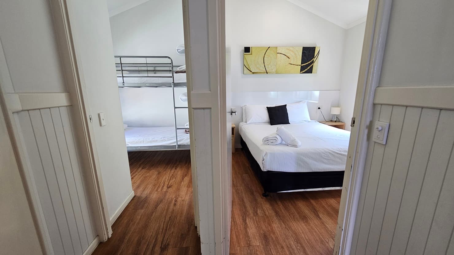 Two doors lead to the two bedrooms. The bedroom on the right features a queen bed with white covers, arranged with pillows and rolled towels. A bedside lamp sits on the right, and a framed decoration is placed above the bed. To the left is a room with a bunk bed covered in white linens. The floor is timber, and the pitched ceiling and walls are all white.