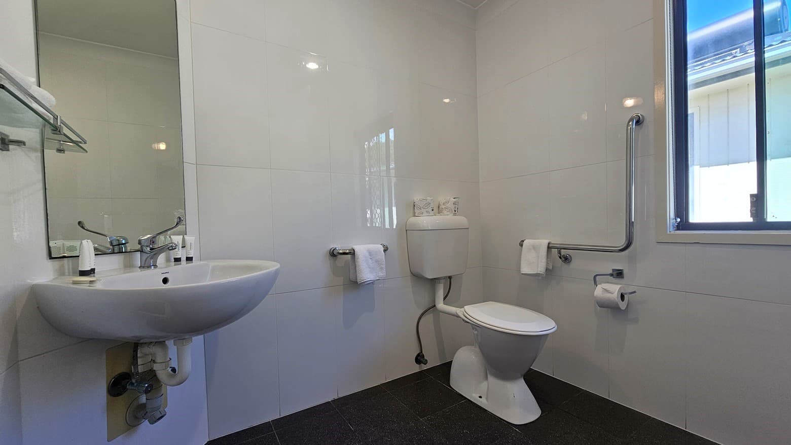 The accessible bathroom features a toilet with hand grabs positioned on the right. A glass window is on the right, adjacent to a hand basin and a wall-mounted mirror. The floor has grey tiles, and the walls are tiled with white tiles. A towel holder is mounted to the wall on the left.