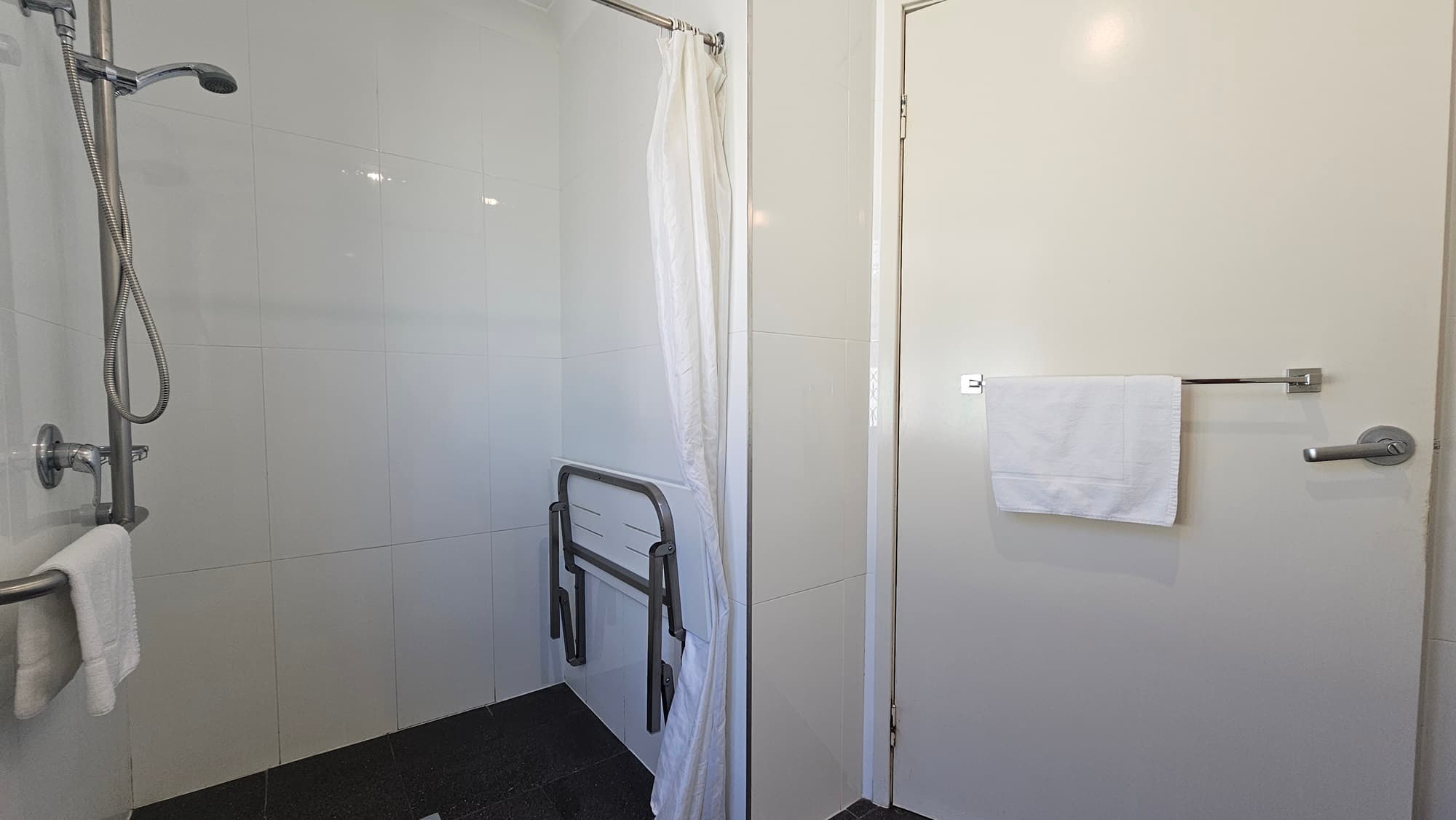 The accessible bathroom features a shower with curtains and hand grabs. A foldable bench is positioned to the right.  A towel holder is mounted to the wall on the left. To the right is a door with a towel holder. The floor has grey tiles, and the walls are tiled with white tiles.
