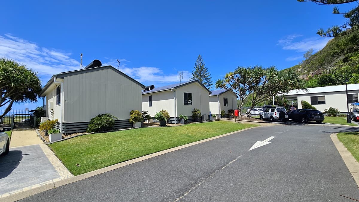 A bitumen road with markings. On the left side of the road, there is a white low set building with a dark grey roof, and windows visible on the side. In front of the building, there is a well-manicured lawn with a small garden bed hosting several plants. On the right side of the road, there is a similar white building featuring a grey roof, with its facade and entrance door visible. There are also a parked black car and a street lamp on the right side near the second building.