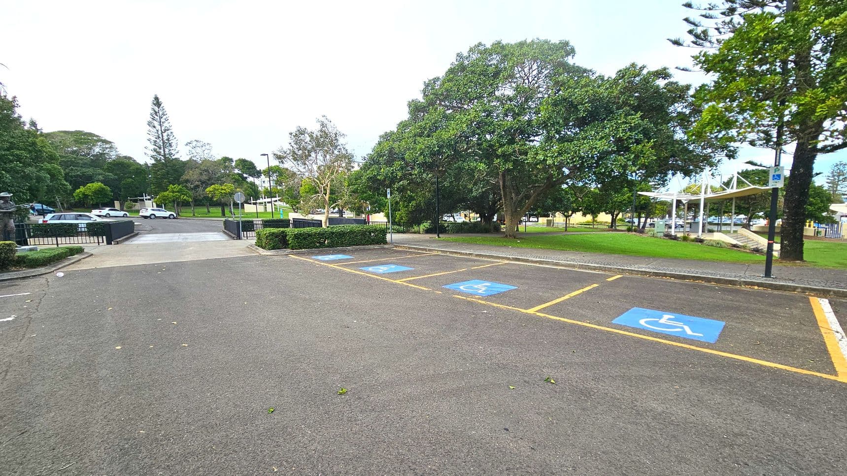 A parking area with several marked disabled parking spaces, indicated by blue paint and wheelchair symbols on the asphalt. The area is bordered by a low hedge and trees, providing a natural boundary. A paved path leads from the parking area towards a grassy park with a white canopy structure visible in the distance. The space feels open with clear transitions from asphalt to grass and paved paths.