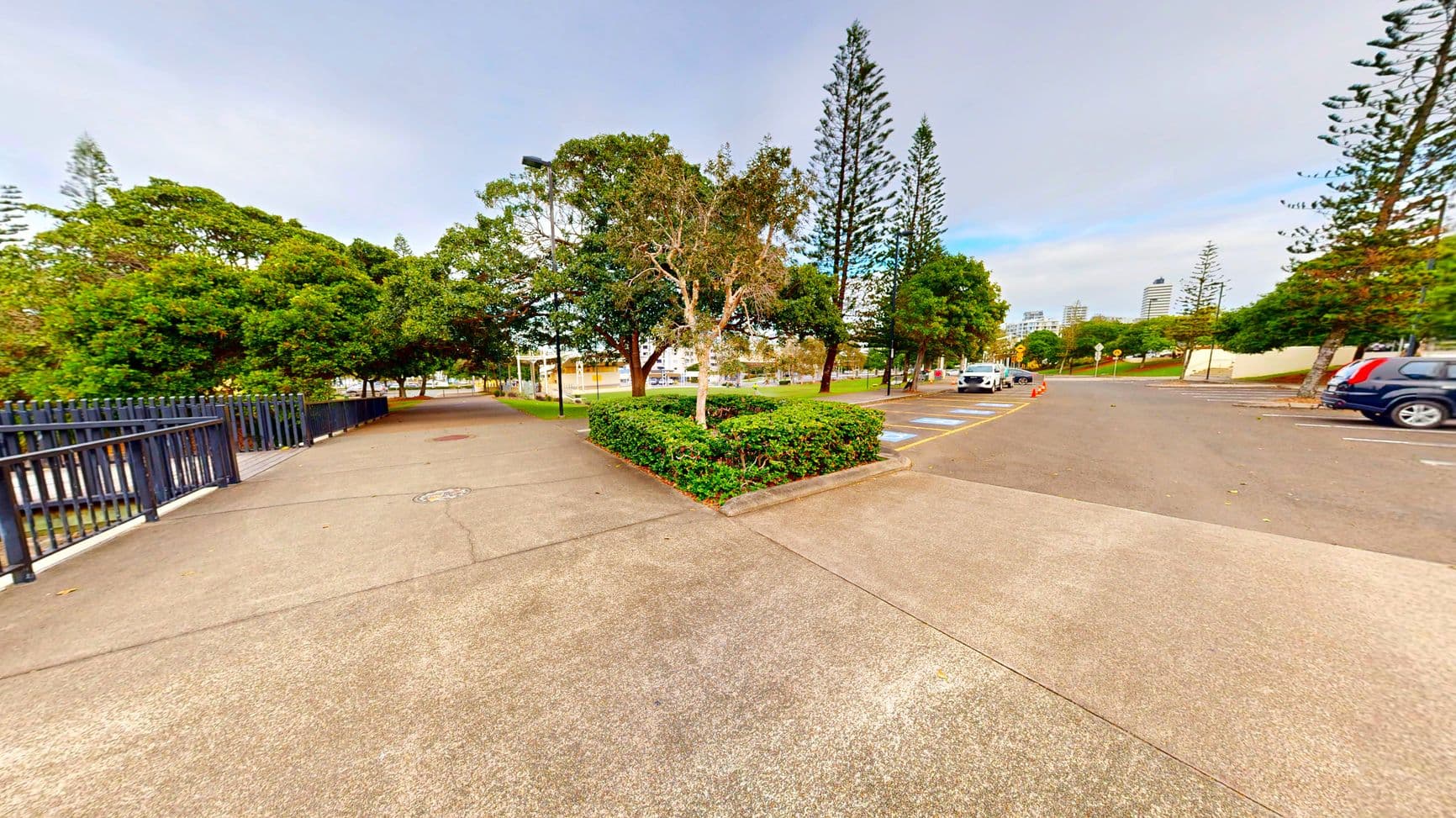 A paved area with a T-junction path bordered by a low hedge and trees. To the left, a black metal fence runs parallel to the path. On the right, a car park with marked spaces and several parked cars is visible. Tall trees and a few buildings are in the background. The area feels open with clear pathways and a mix of concrete and asphalt surfaces.