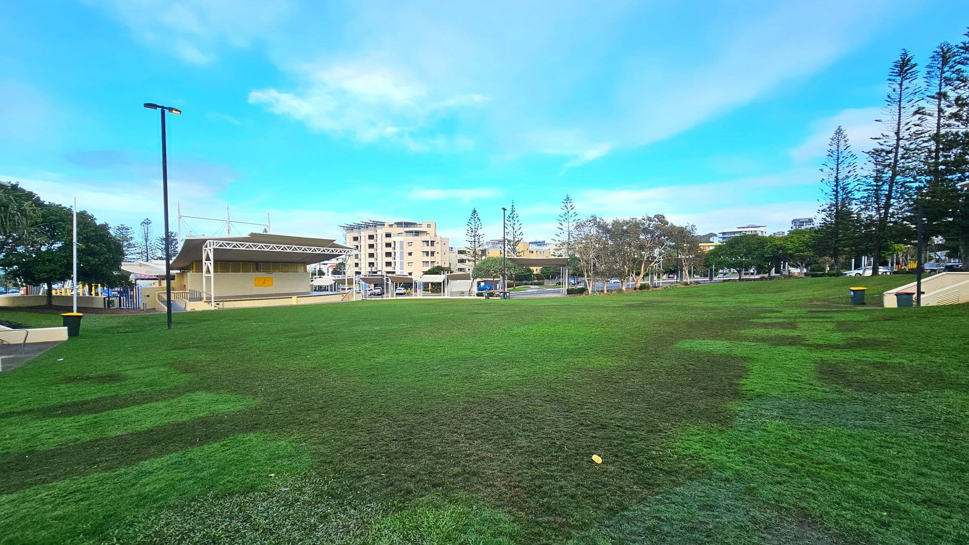 A spacious grassy park area with a gently sloping lawn. To the left, a small stage structure with a geometric roof and yellow walls is visible. Several tall pine trees line the background, alongside modern multi-storey buildings. A few scattered benches and bins are present, and a tall streetlamp stands prominently. The area feels open with no visible barriers, and the grass transitions smoothly to paved paths.