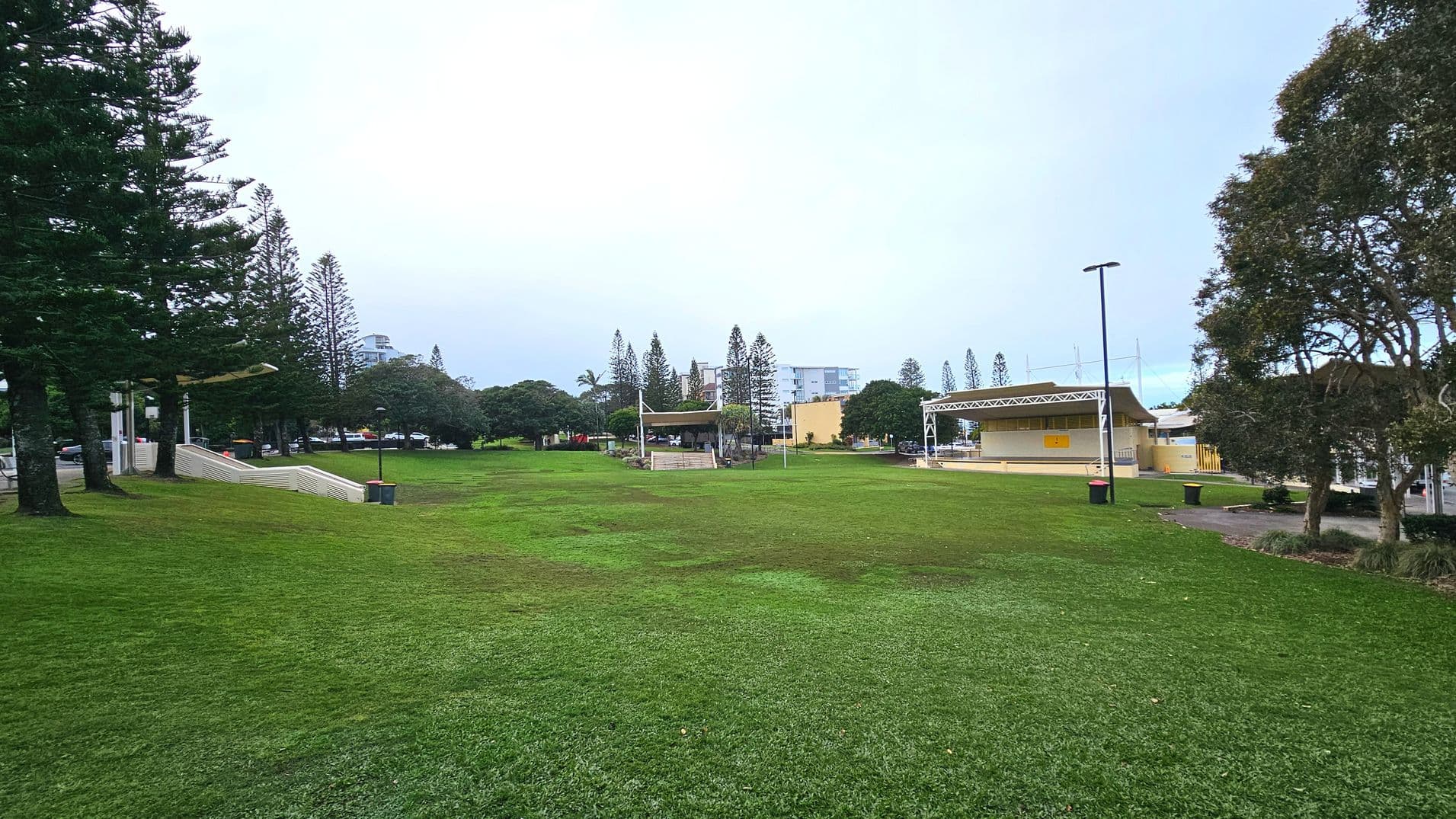 A spacious grassy park area with a gently sloping lawn. On the right, a modern pavilion with a flat roof and open sides is visible, adjacent to a paved path. To the left, a series of wide, white steps lead down from a higher level. Tall trees line the left side, providing a natural boundary. Several buildings are visible in the background, adding an urban touch to the otherwise open space.