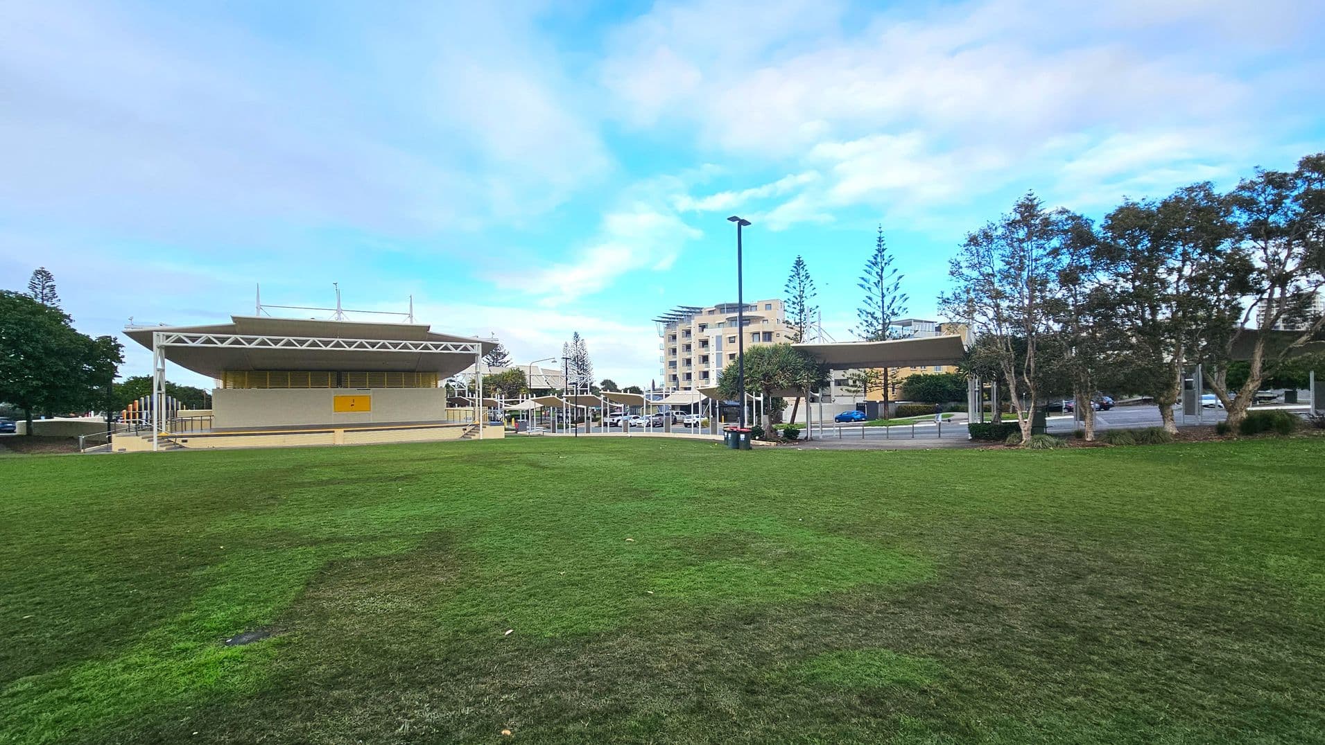 A grassy open area leads to a modern building with a flat, overhanging roof supported by white beams. The building has a yellow panel on its facade and is surrounded by a paved area with railings. To the right, several trees line the edge of the grass, and a street with parked cars is visible. In the background, a multi-storey building rises, adding an urban element to the scene.