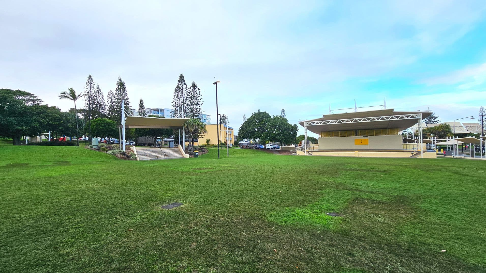 A grassy open area with two prominent structures. On the left, a covered stage with steps leading up, surrounded by trees and a few people. On the right, a larger building with a geometric roof and a yellow sign on its wall. Both structures are set against a backdrop of tall trees and distant buildings. The ground is mostly grass with some patches of dirt, and a tall lamp post stands in the centre.