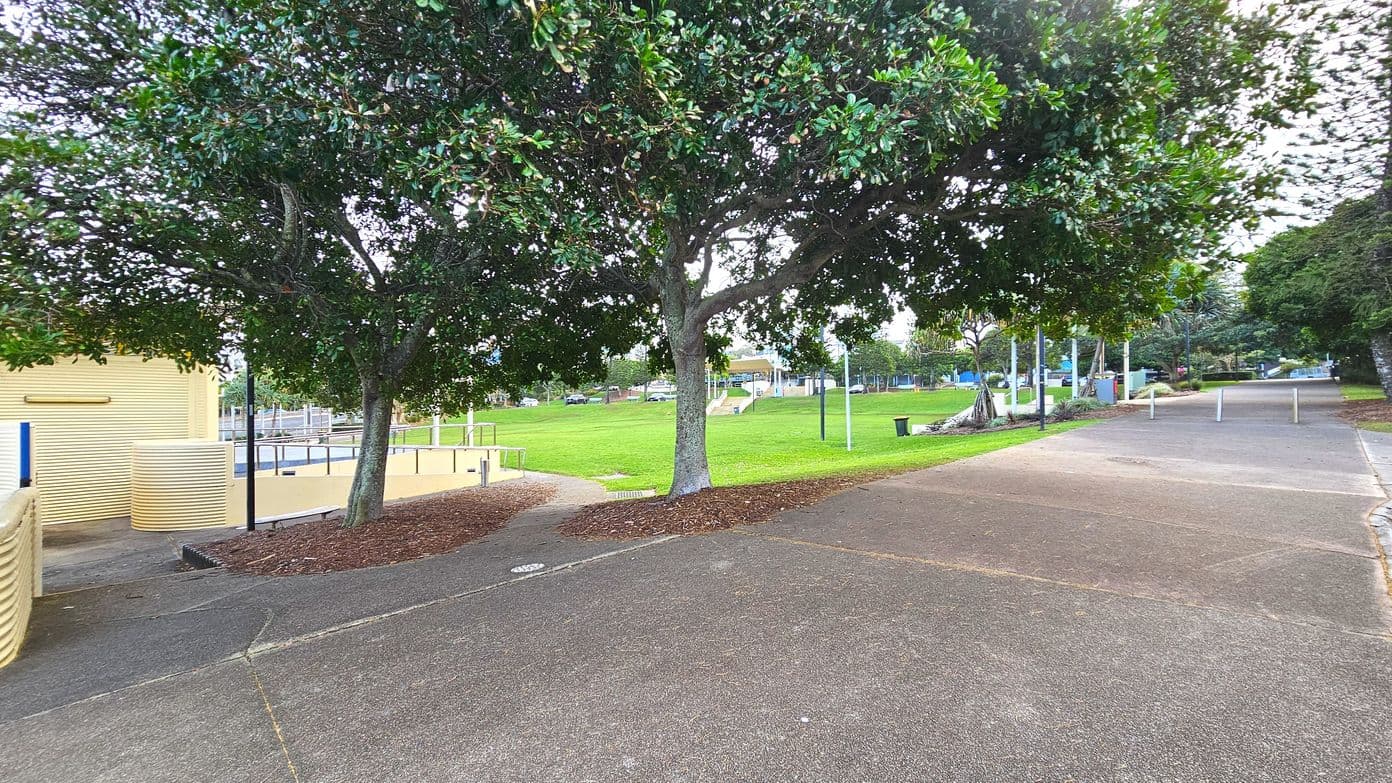 A park scene with a paved pathway curving around a large tree in the centre. To the left, a yellow building with ribbed walls and a ramp is visible. The path transitions from concrete to a reddish-brown surface. Beyond the tree, a grassy area extends with scattered trees and a few poles. The space feels open with no visible barriers. A few bollards line the path on the right, indicating pedestrian access.