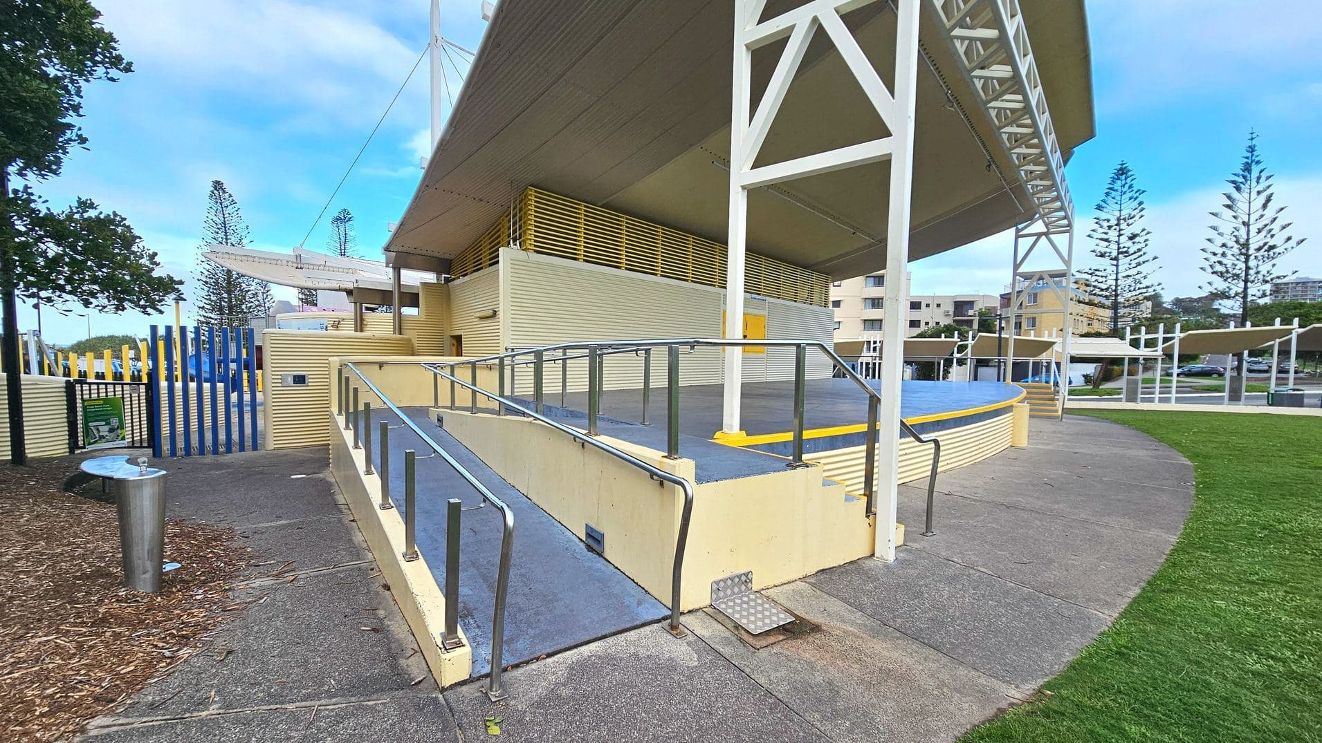 A modern outdoor amphitheatre with a large, angular roof supported by white beams. The structure features a blue stage with yellow accents and a surrounding yellow railing. A ramp with metal handrails leads up to the stage, adjacent to a set of steps. The ground is a mix of concrete and grass, with a metal drinking fountain nearby. Blue vertical bars form a fence on the left, and trees and buildings are visible in the background.