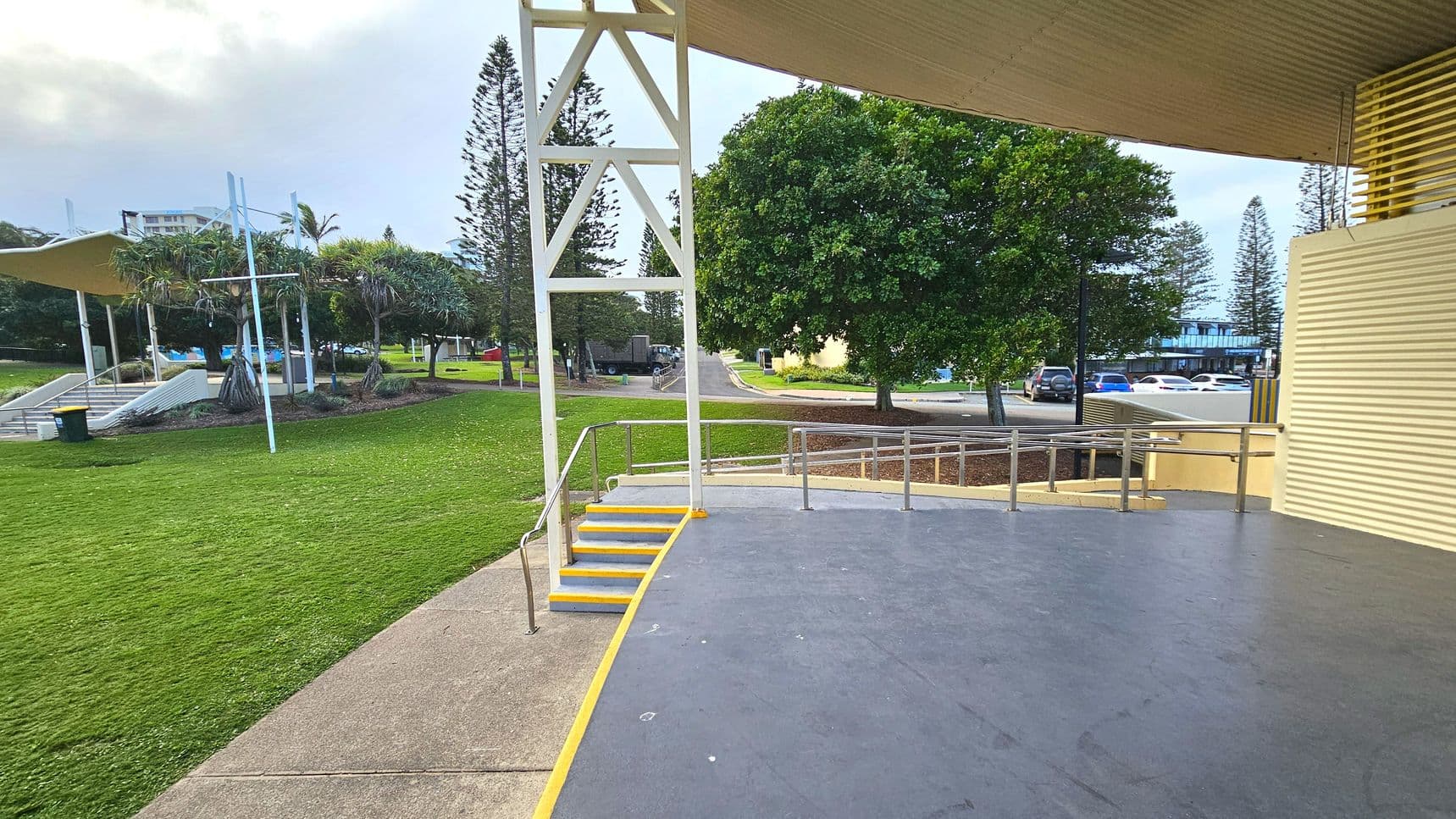 An outdoor stage area with a grey floor and yellow-edged steps leading down to a concrete path. A metal railing lines the steps and the stage edge. To the left, a grassy area with palm trees and a covered seating area is visible. In the background, a road with parked cars and a large tree can be seen. The stage is partially enclosed by a corrugated metal wall and supported by a white metal framework.
