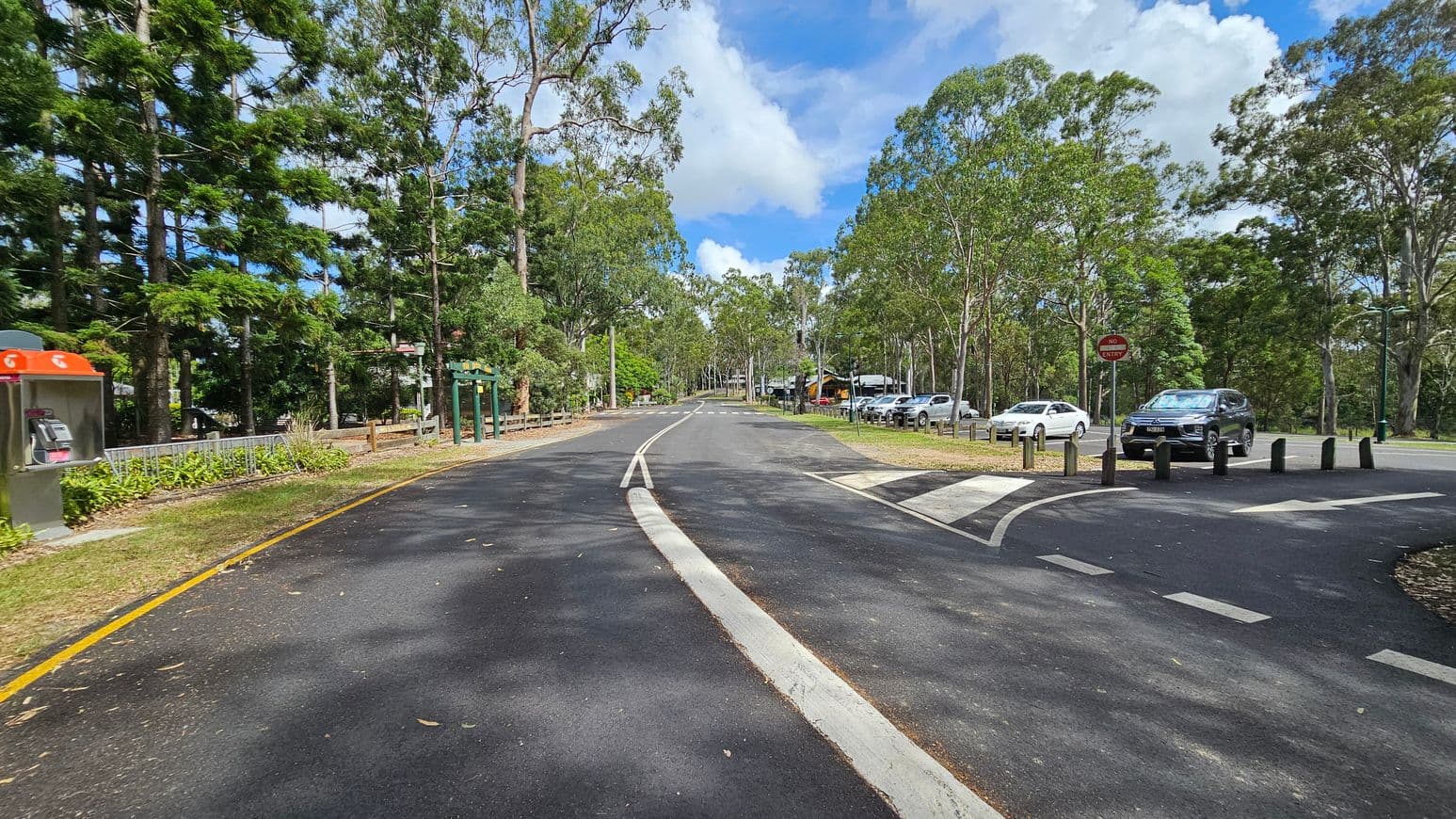 A road curves gently to the left, bordered by tall trees on both sides. On the left, there's a phone booth with an orange top and a green information board. On the right, several parked cars are visible near a "No Entry" sign. The road surface is dark with white lane markings, and there are bollards separating the road from a pedestrian crossing. The area is surrounded by dense greenery, creating a natural setting.