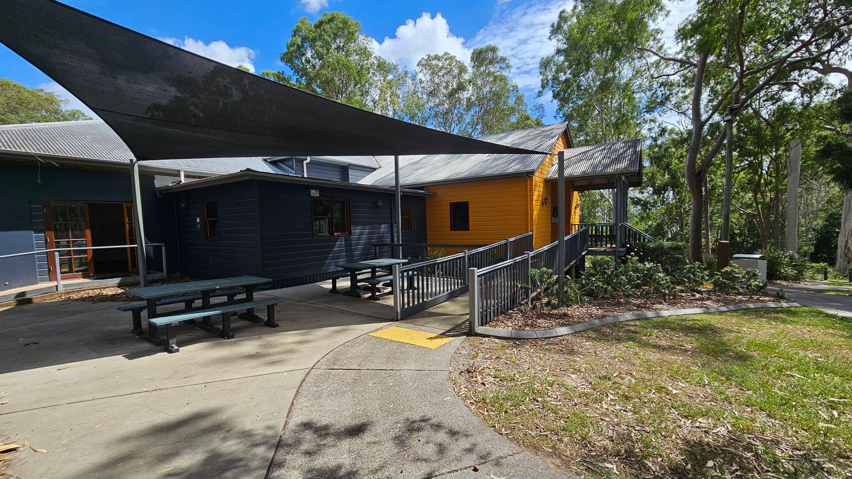 A building with a corrugated metal roof and a combination of dark and bright yellow walls. In front, there are two picnic tables under a large triangular shade sail. A ramp with metal railings leads to the entrance. The ground is a mix of concrete and green-painted surfaces, with a small garden area to the right. Trees surround the area, providing a natural backdrop.
