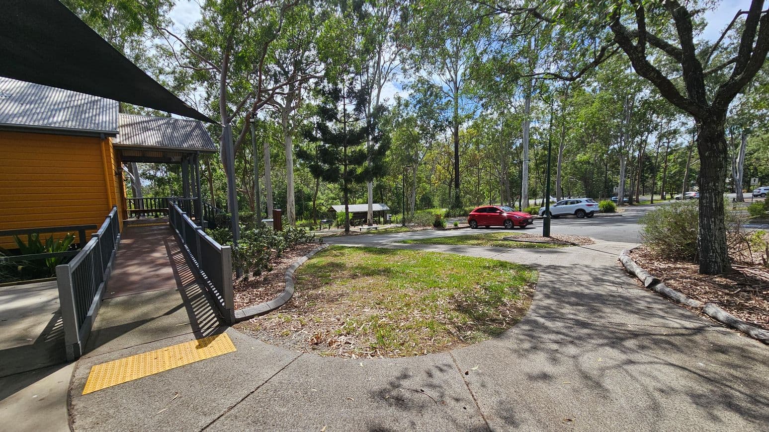 A pathway leads from the foreground to a yellow building on the left, featuring a corrugated metal roof and a ramp with railings. In the middle, there is a grassy area with scattered leaves and a few trees. On the right, a car park is visible with a red car and a white car parked. The background is filled with tall trees, and a small shelter with a picnic table is partially visible. The ground is a mix of concrete and grass, with some mulch around the trees.