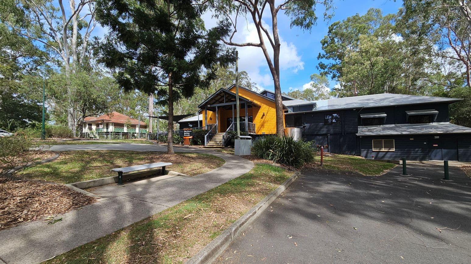 A pathway curves through a park-like setting with a bench on the left. In the centre, a yellow wooden building with a gabled roof and steps leading to the entrance is surrounded by trees. To the right, a dark-coloured building with a sloped roof is partially visible. The ground is covered with leaves and grass, and the area is bordered by a paved road. Tall trees with dense foliage are in the background.
