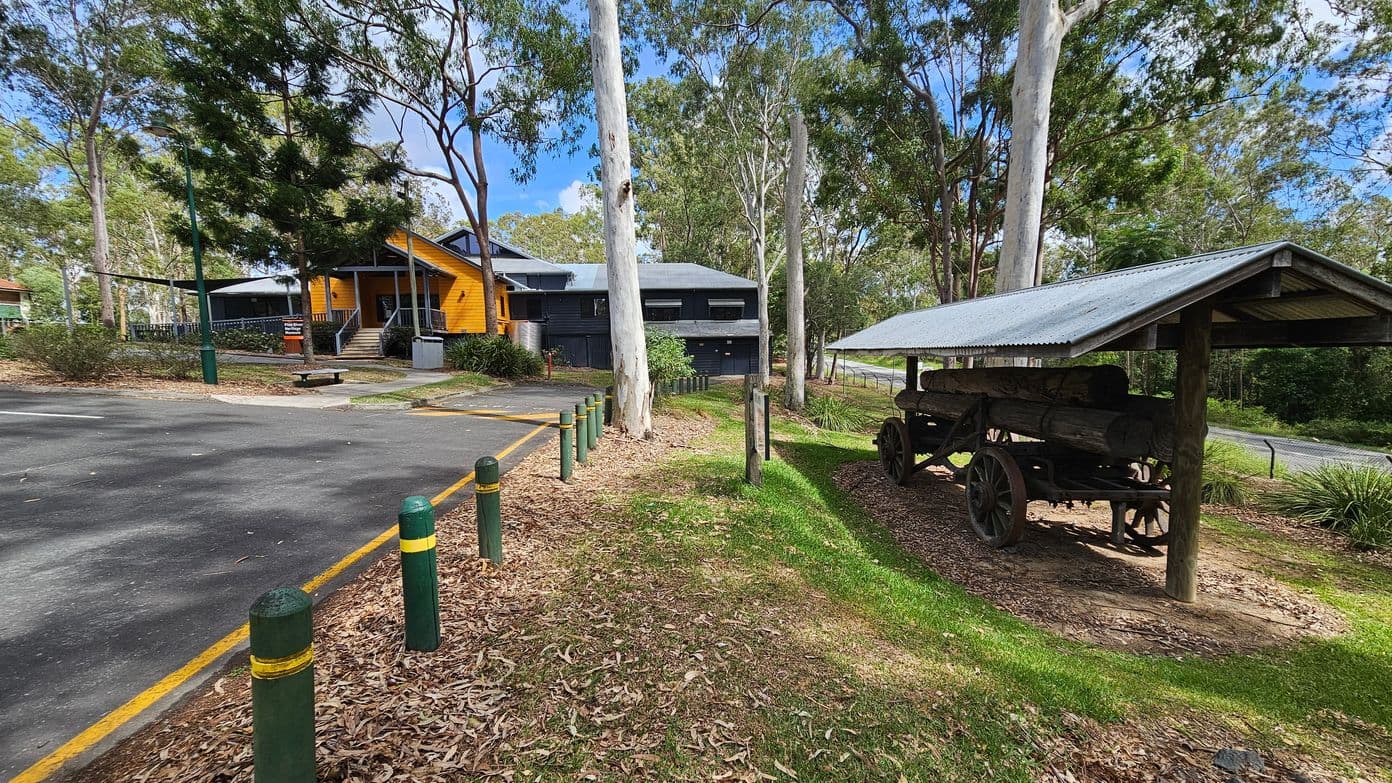 A rustic scene with a yellow wooden building on the left, featuring a grey roof and a small staircase leading to the entrance. In front, there's a paved area with green bollards and a bench. On the right, an old wooden cart with large wheels is sheltered under a corrugated metal roof. The ground is covered with grass and fallen leaves. Tall trees surround the area, providing a natural backdrop.