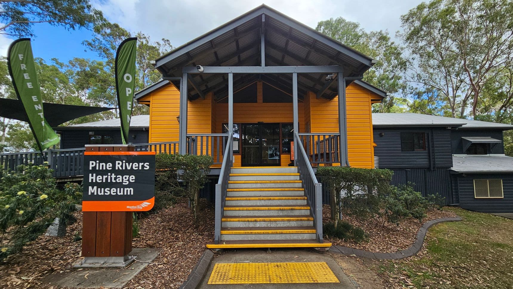 A building with a bright yellow facade and a grey roof, featuring a central entrance with steps leading up to it. On the left, two green flags display the words "FREE ENTRY" and "OPEN". A sign in front reads "Pine Rivers Heritage Museum" with "Moreton Bay" below. The surrounding area has trees and bushes, with a path leading to the entrance. The building has a veranda with grey railings. The ground is covered with mulch and grass.