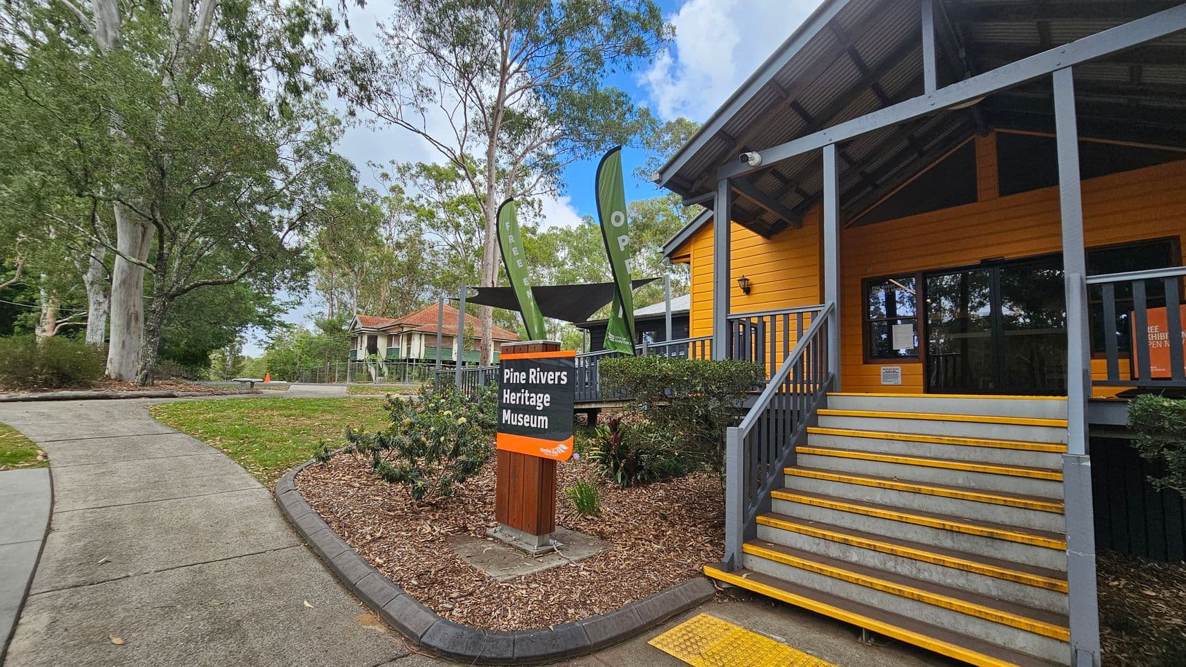 A yellow building with a grey roof and steps leading up to a glass entrance is on the right. A sign in front reads "Pine Rivers Heritage Museum." Two green flags with "OPEN" are near the entrance. On the left, a path curves through a grassy area with large trees. Another building with a red roof is visible in the background. The ground is covered with mulch and bordered by low shrubs.