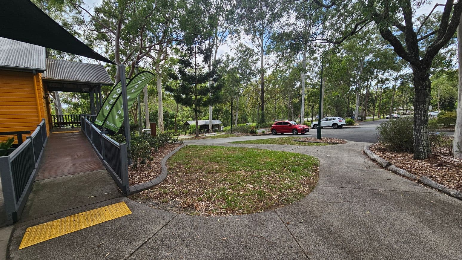 A pathway curves through a park-like setting. On the left, a wooden building with a corrugated metal roof has a ramp leading to it, flanked by green flags. In the centre, a grassy area is bordered by mulch and trees. To the right, a red car and a white car are parked on a paved road. Tall trees with slender trunks are scattered throughout, providing a canopy of leaves. The ground is a mix of concrete, grass, and mulch.