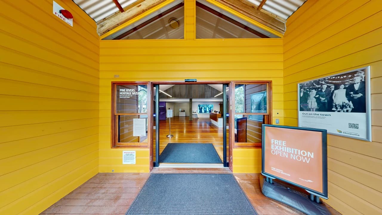 Entrance to a building with bright yellow wooden walls. In the centre, double glass doors lead into an interior with wooden flooring. Above the doors, a triangular window is visible. On the right, a large sign reads "Free Exhibition Open Now" and a black-and-white photo is displayed. The ceiling is sloped with exposed beams. A mat is placed on the floor at the entrance.