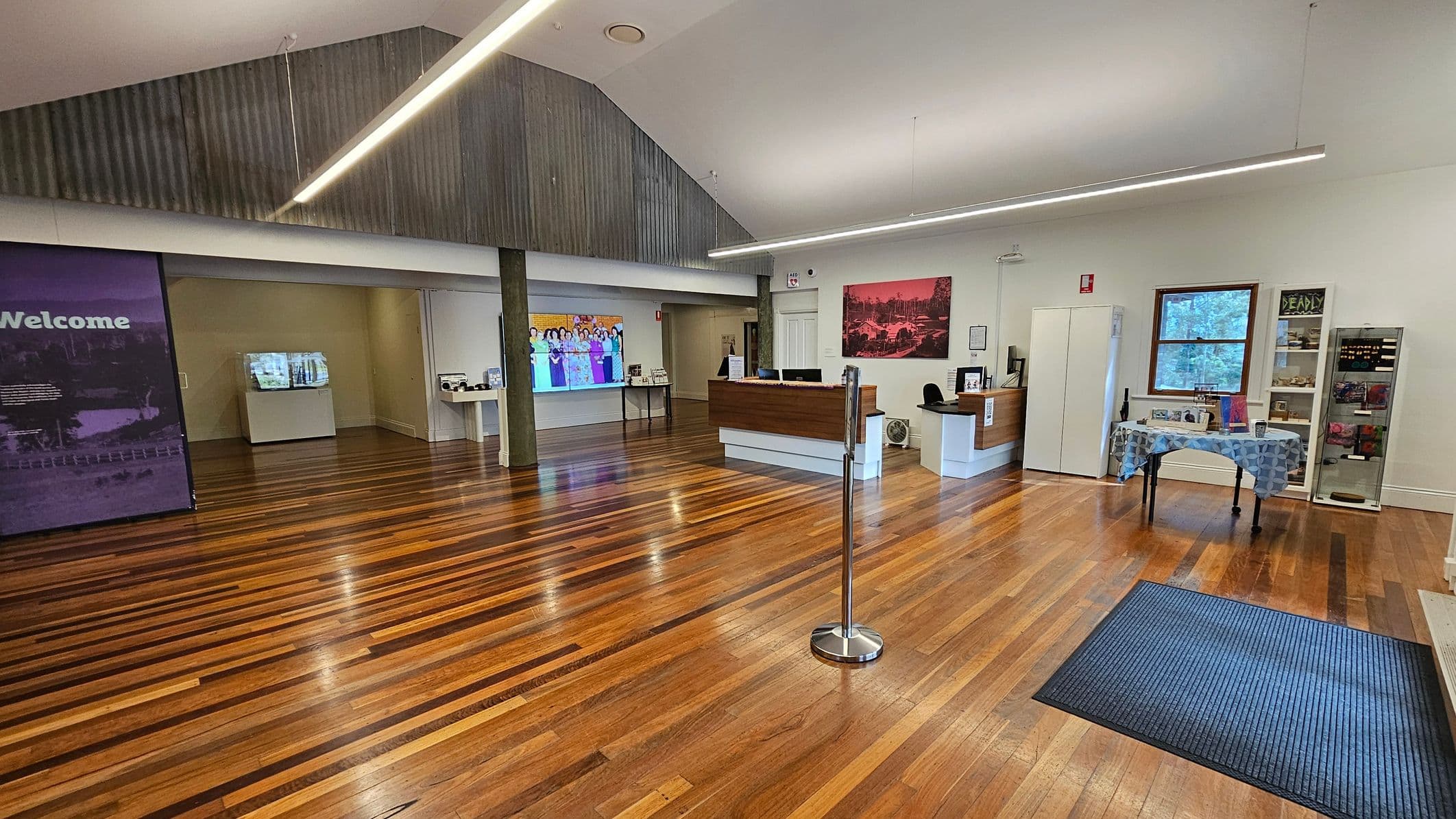 A spacious room with wooden flooring and a high ceiling. On the left, a large purple sign with "Welcome" is visible. In the centre, a reception desk with a computer and a chair is positioned against the wall, with a colourful image displayed on a screen behind it. On the right, a window with a view of trees, a table with a patterned cloth, and a display cabinet are present. A blue mat is on the floor near the entrance. The ceiling features long, linear lights.