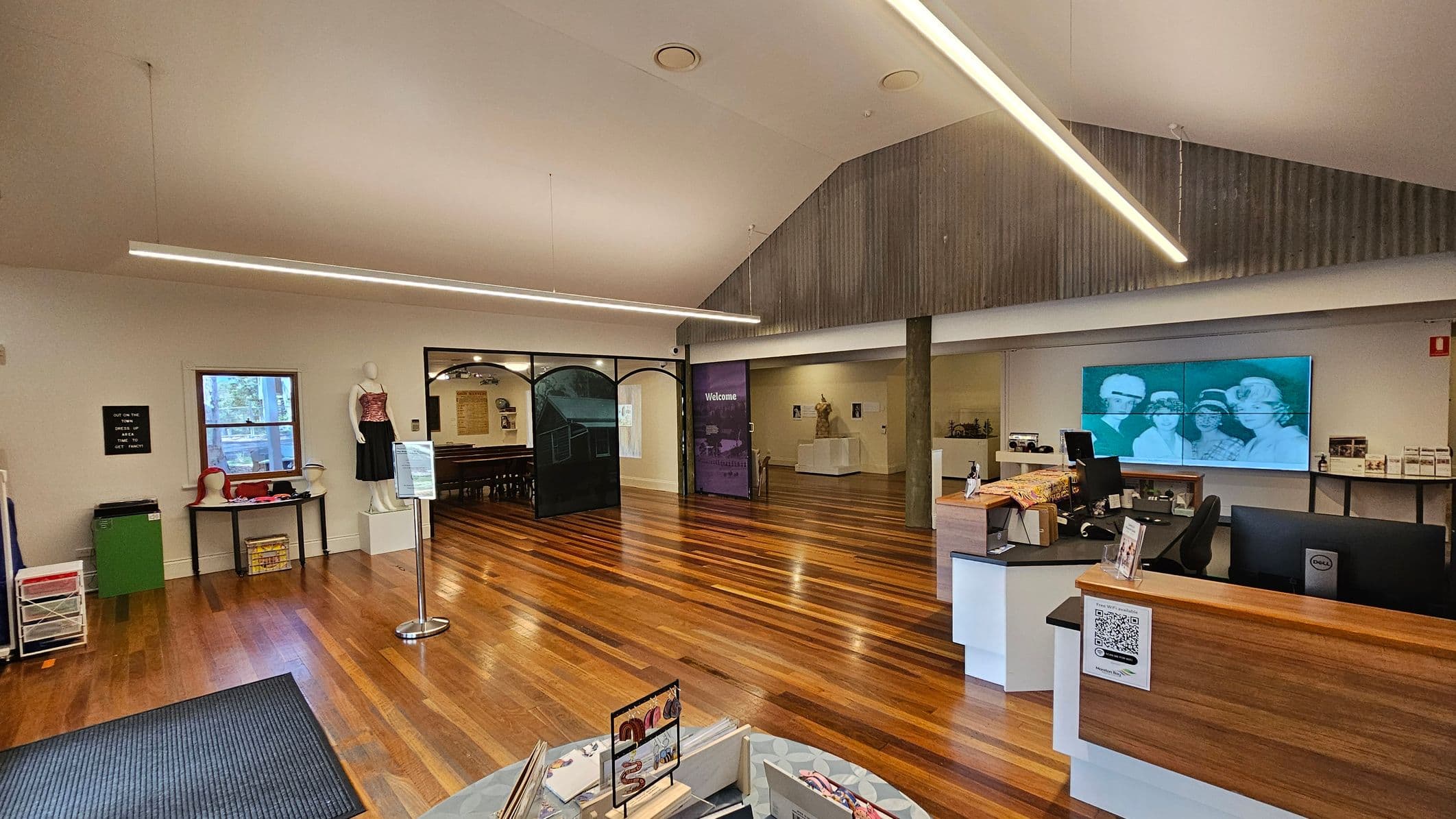 A spacious room with wooden flooring and a high ceiling. On the left, a mannequin dressed in a red and black outfit stands near a window, with a table displaying hats and other items. In the centre, a partition with "Welcome" written on it leads to another area. On the right, a reception desk with a computer and a QR code is in front of a wall displaying a large black-and-white photograph. The ceiling features long, linear lights.