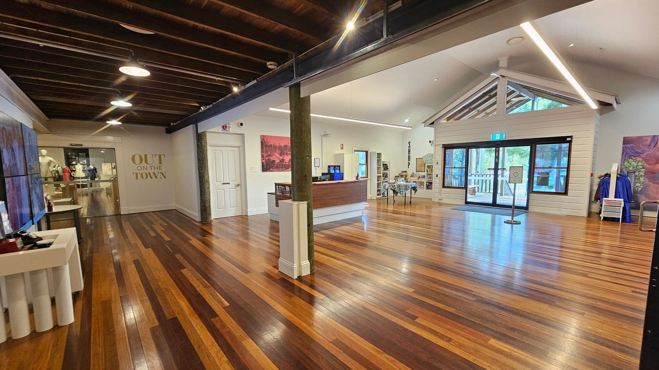 A spacious interior with polished wooden floors and a high ceiling. On the left, there's a display area with mannequins and a sign reading "OUT ON THE TOWN." In the centre, a wooden reception desk is visible, with a small shop area behind it. On the right, a structure resembling a small house with large windows and a door leading outside. The ceiling features exposed beams and modern lighting.