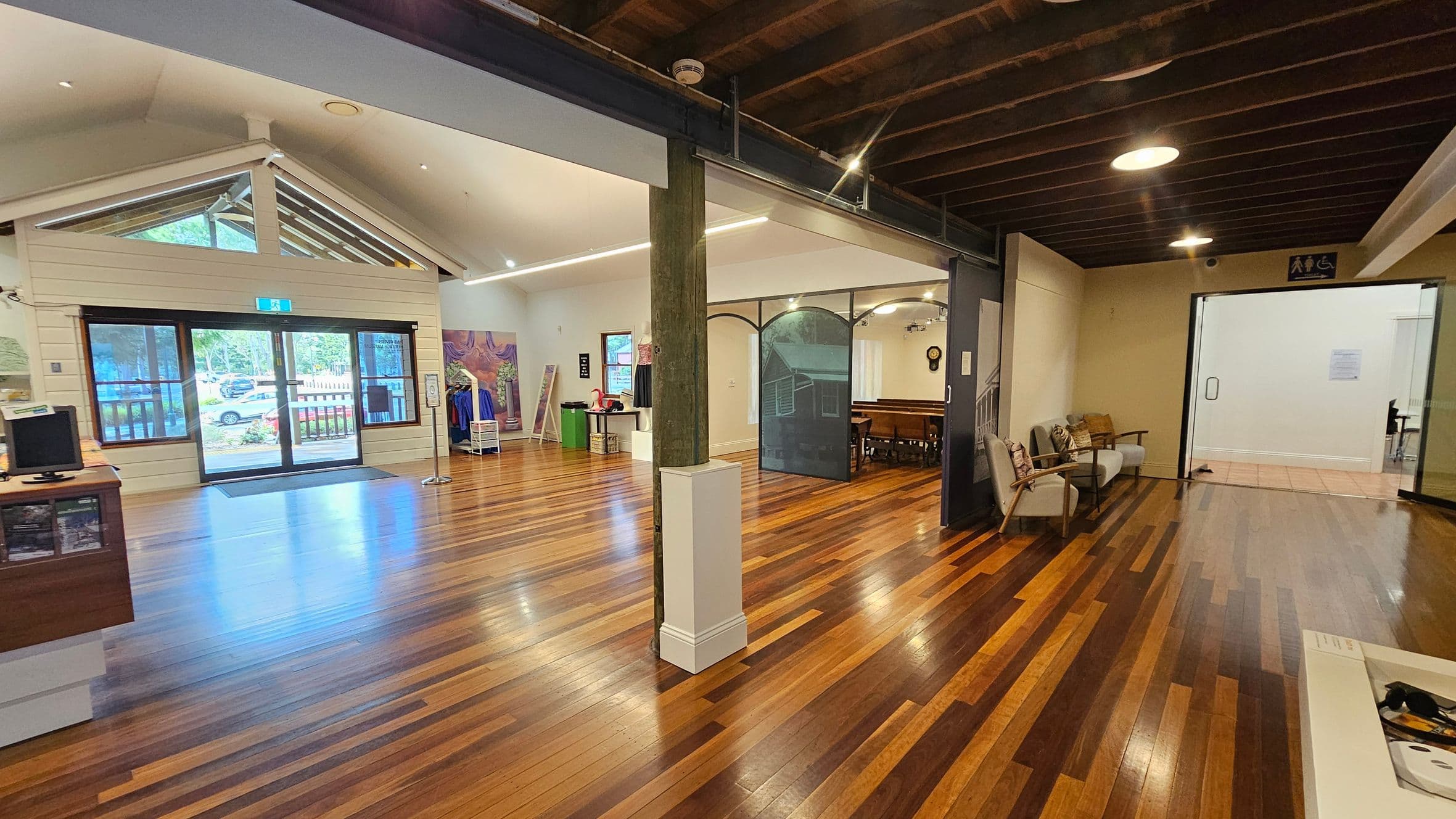 A spacious interior with wooden flooring and a high ceiling. On the left, there is a large glass door with a triangular window above, leading outside. A counter with a computer is nearby. In the centre, a wooden pillar divides the space, with a display area featuring clothing and artwork. On the right, there are chairs against the wall and an open doorway leading to another room. The ceiling has exposed beams and recessed lighting.