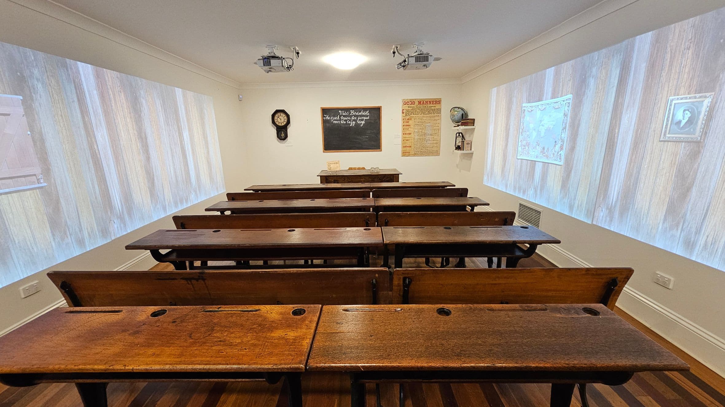 A vintage classroom with wooden desks arranged in rows. At the front, a blackboard displays writing, flanked by a wall clock and a poster titled "Good Manners". A teacher's desk is positioned beneath the blackboard. The side walls have projections of wooden panels, a map, and a framed portrait. The ceiling features two projectors and a central light. The floor is wooden, complementing the desks.