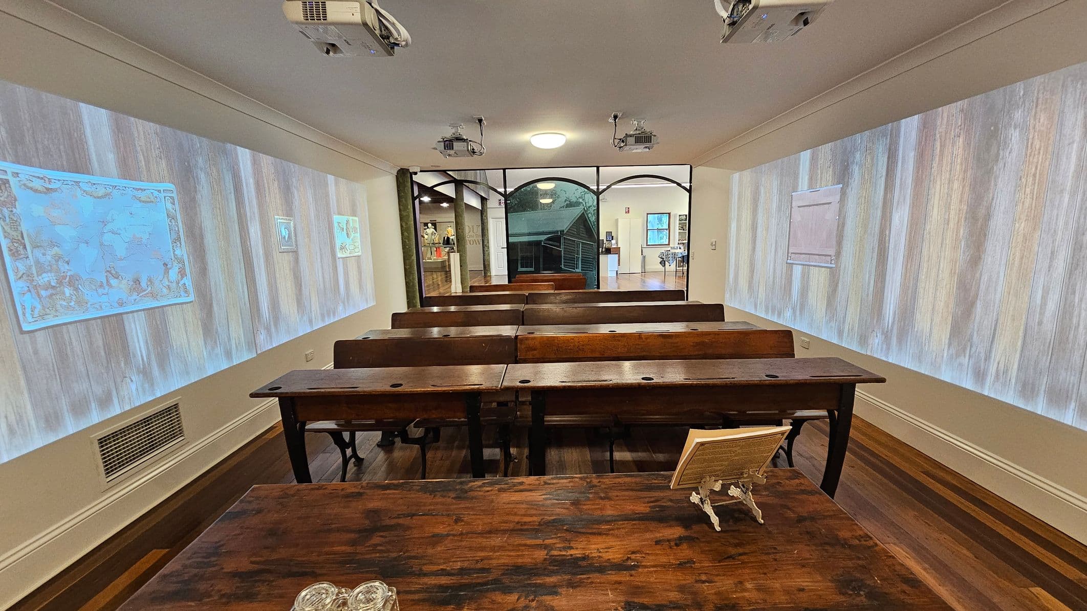A classroom with wooden desks and benches arranged in rows. The walls on both sides display projected images, including maps. The ceiling has several projectors and a central light fixture. At the back, a glass partition reveals another room with displays and a staircase. The floor is wooden, matching the desks.
