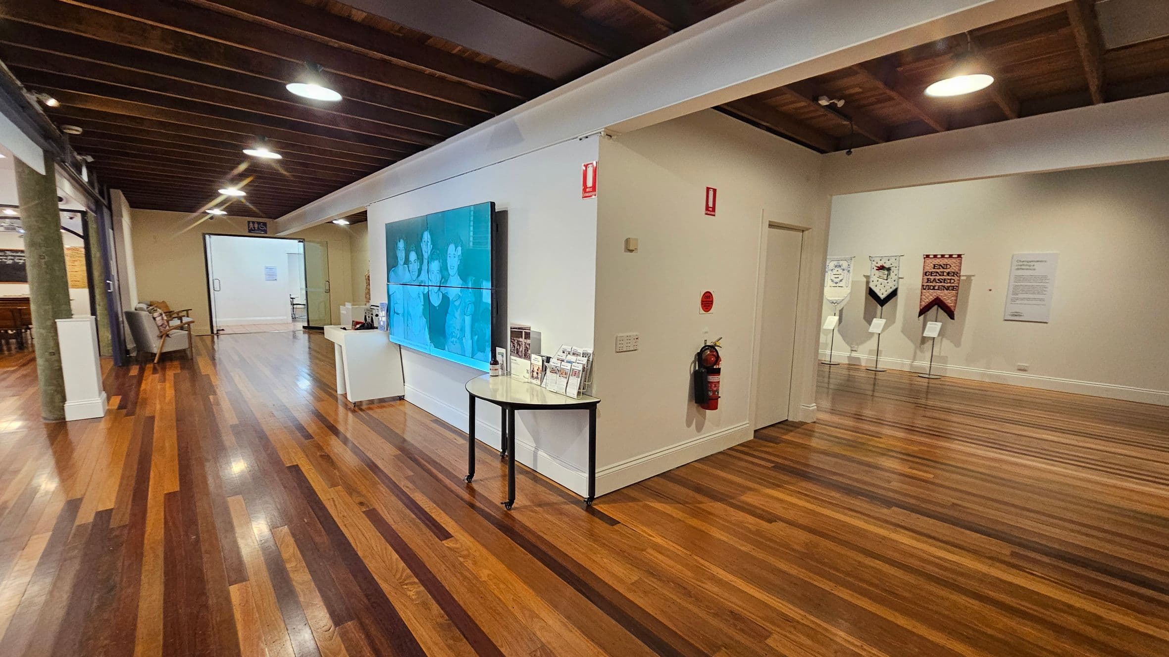 A spacious room with polished wooden floors and a wooden beam ceiling. On the left, there is a seating area with chairs and a table. In the centre, a large screen displays an image, mounted on a white wall with a small table below it holding brochures. On the right, banners with text are displayed on stands against the wall. A fire extinguisher is mounted on the wall near a door. The room is well-lit with ceiling lights.