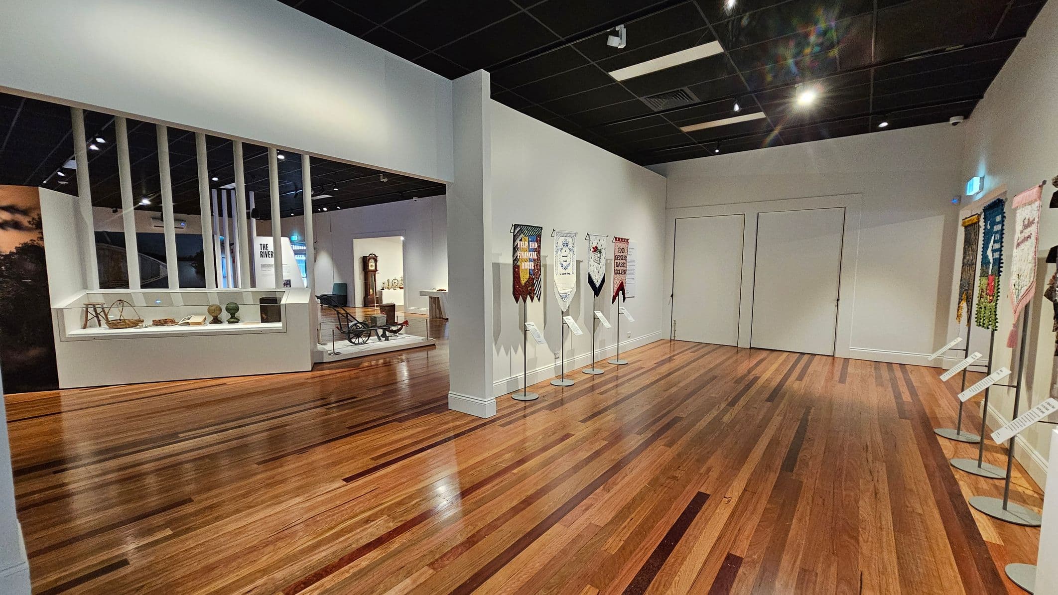 A museum interior with wooden flooring and a black ceiling. On the right, several banners are displayed on stands against a light-coloured wall. In the centre, a doorway leads to another exhibit area with a grandfather clock and various artefacts. On the left, a glass display case contains historical items, and vertical white beams separate this section from the rest of the room.