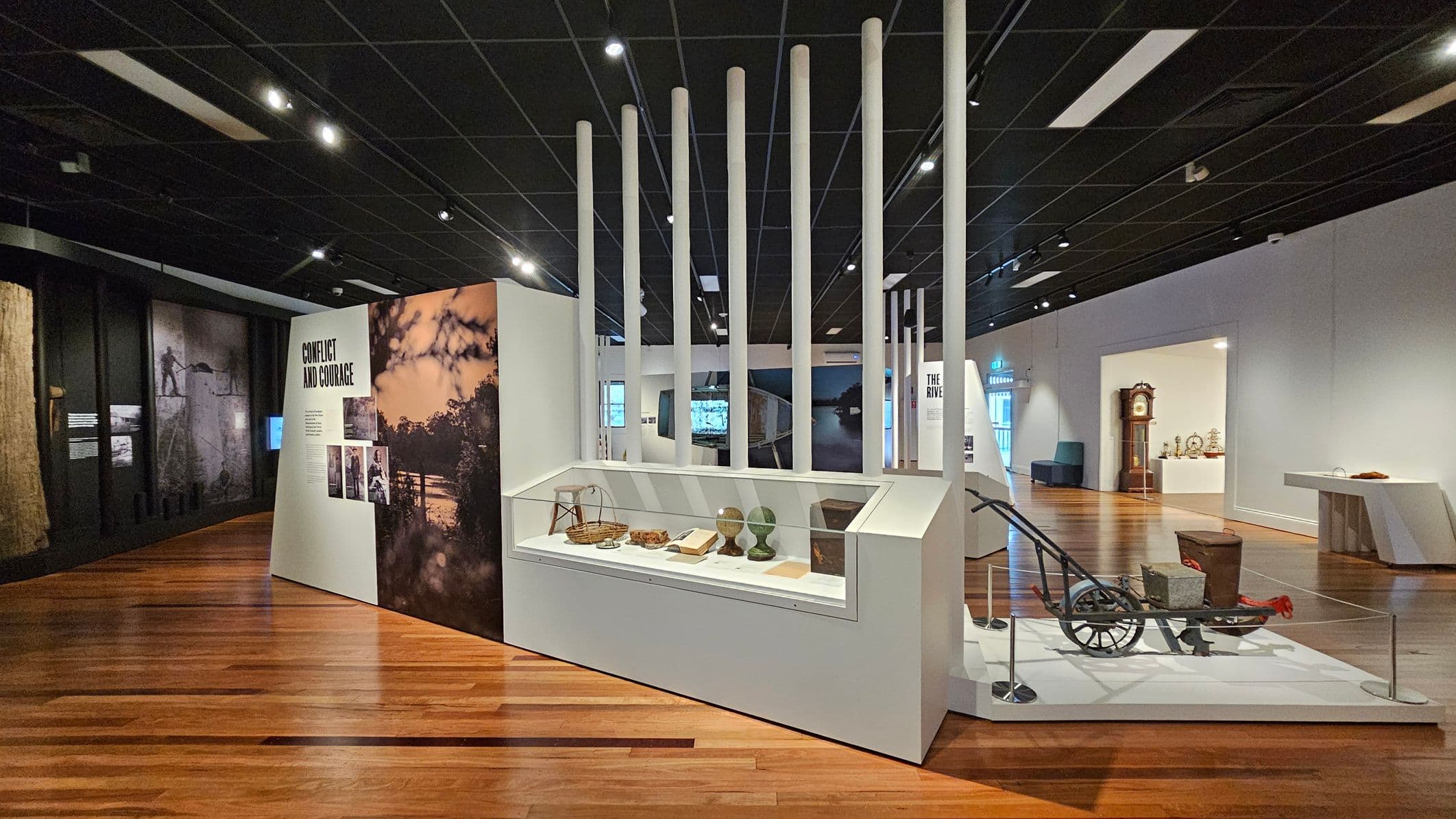 A museum exhibit with a wooden floor and a dark ceiling. On the left, a display titled "Conflict and Courage" features images and text. In the centre, a glass case contains various artefacts, including baskets and books, with tall white poles behind it. On the right, a vintage plough is displayed on a platform, and further back, a grandfather clock and other items are visible in an adjacent room.