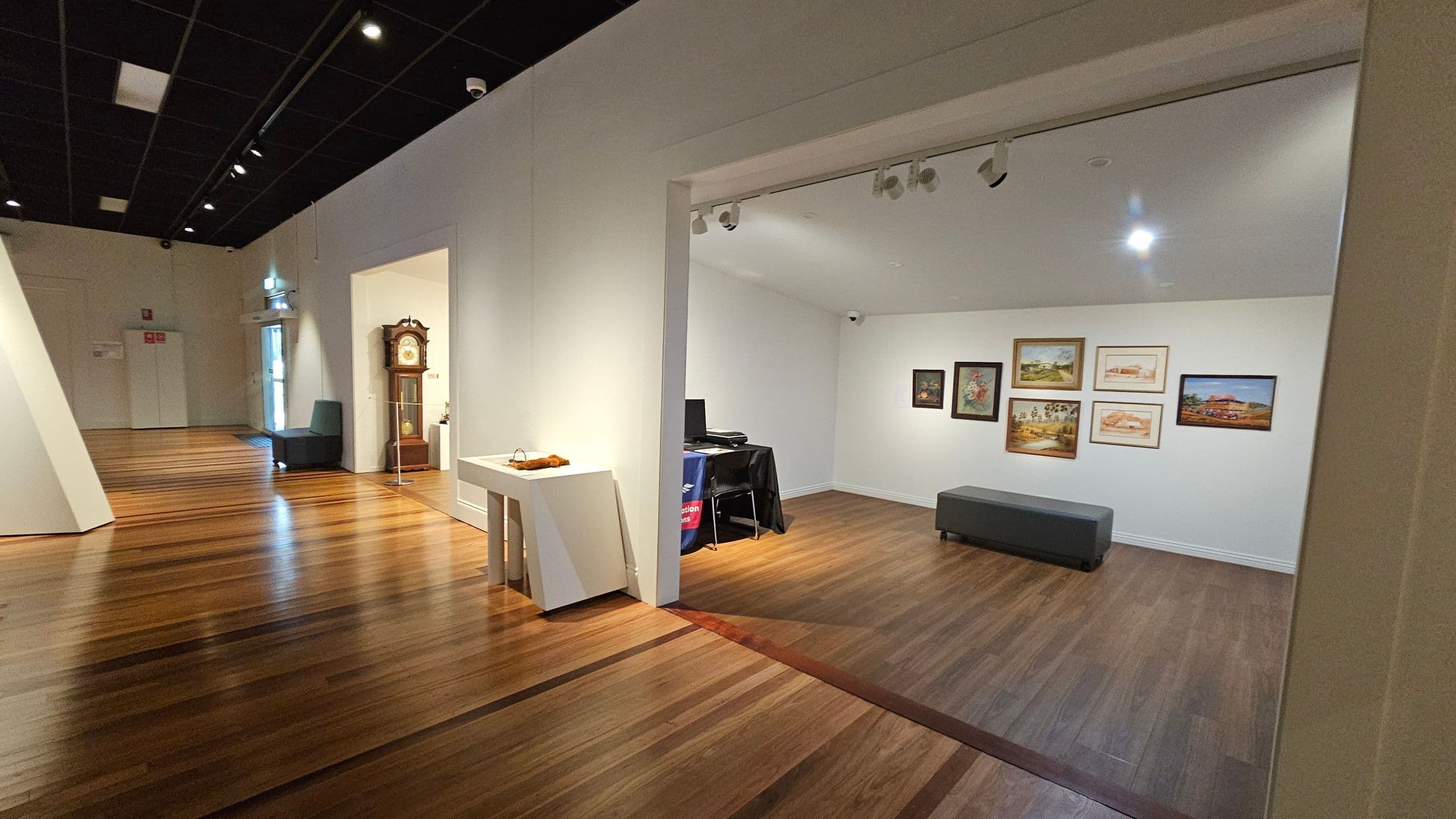A gallery space with wooden flooring and a black ceiling. On the left, there's a hallway with a grandfather clock and a dark chair near a door. In the centre, a small room displays several framed artworks on a white wall, with a grey bench below. A table with a black cloth and computer equipment is in the corner. A white table with decorative items is positioned near the entrance to the room. Track lighting is installed on the ceiling.