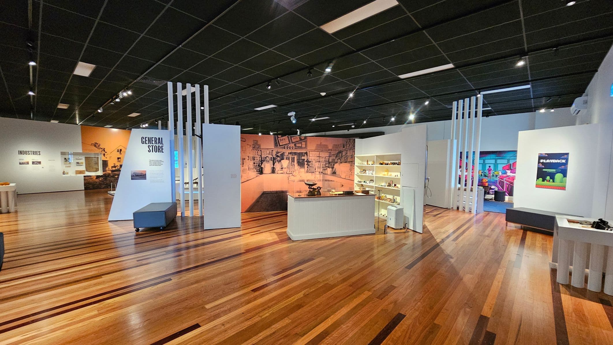 A museum exhibit with wooden flooring and a black tiled ceiling. In the centre, a display labelled "General Store" features a counter with vintage items and shelves filled with small objects. To the left, a wall with "Industries" has various displays. On the right, a poster titled "Playback" is visible. The background includes a mural depicting a historical scene. Bright lighting highlights the exhibits.